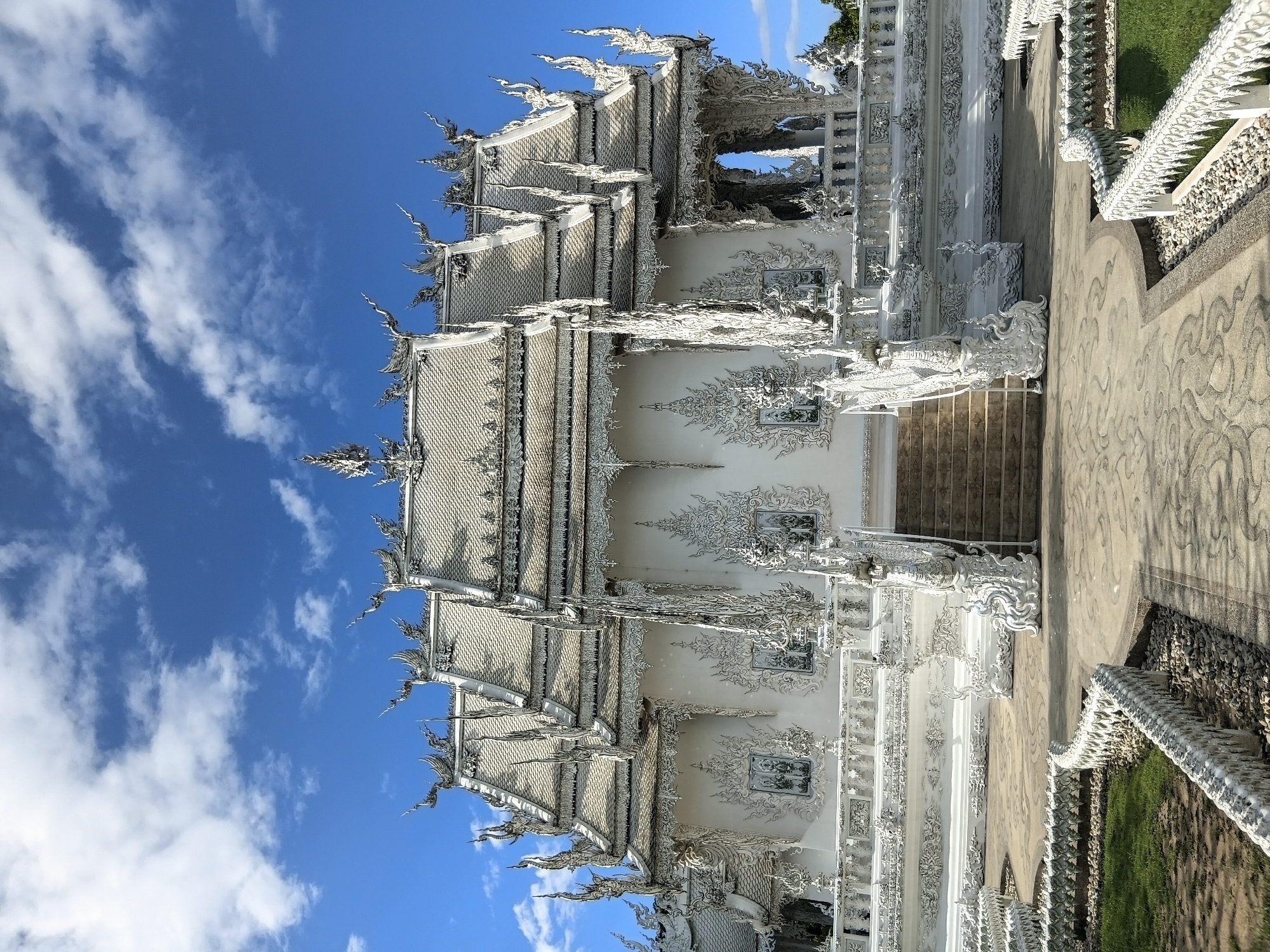 The image shows the Wat Rong Khun, also known as the White Temple, in the city of Ban Rong Khun, Thailand. The temple is a white Buddhist temple with intricate sculptures and details. It is surrounded by a paved courtyard and a lawn. The sky above is blue with white clouds. The temple's architecture is elaborate, featuring ornate roofs, decorative eaves, and detailed carvings on its exterior walls. Staircases with dragon-like railings lead up to the main entrance. The surrounding area is well-maintained with pathways and low white decorative walls that are also sculpted with repetitive figures.