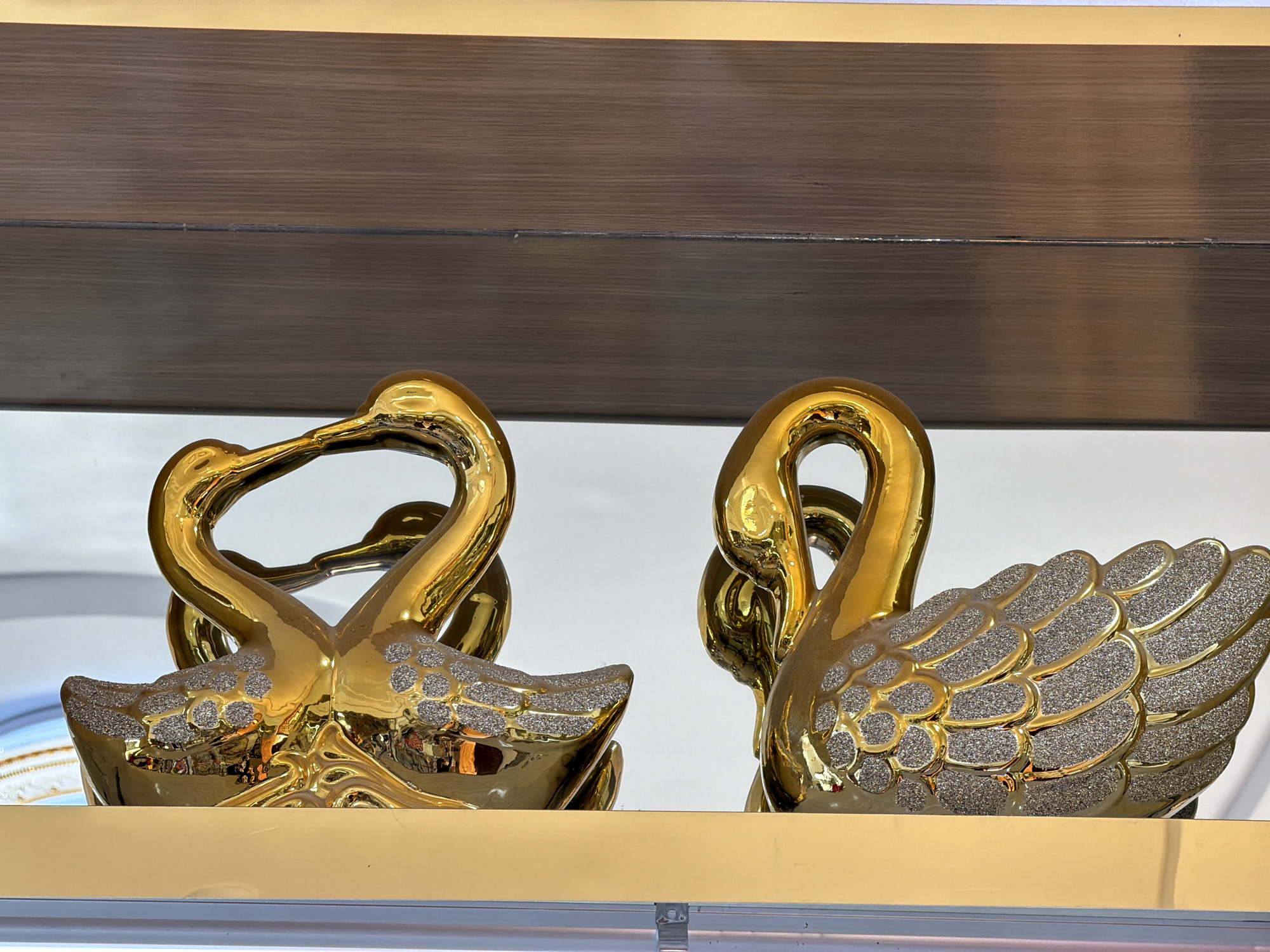 This is a close-up, eye-level, medium shot of two gold swan figurines, likely decorative, displayed on a shelf. The figurine on the left is composed of two intertwined swans forming a heart shape with their necks, while the figurine on the right is a single swan with detailed, glitter-covered wings. The swans have a high-gloss, reflective finish, with the glitter applied to the wings and the back of the head of the left swan. The background consists of a wooden shelf above and a lighter, possibly reflective surface below. The lighting appears to be artificial, creating bright highlights on the golden surfaces. The setting is indoors, on a shelf, and appears to be a retail or display environment. There are no people or animals present. The image does not provide any specific information or context related to Zangiata, Uzbekistan.