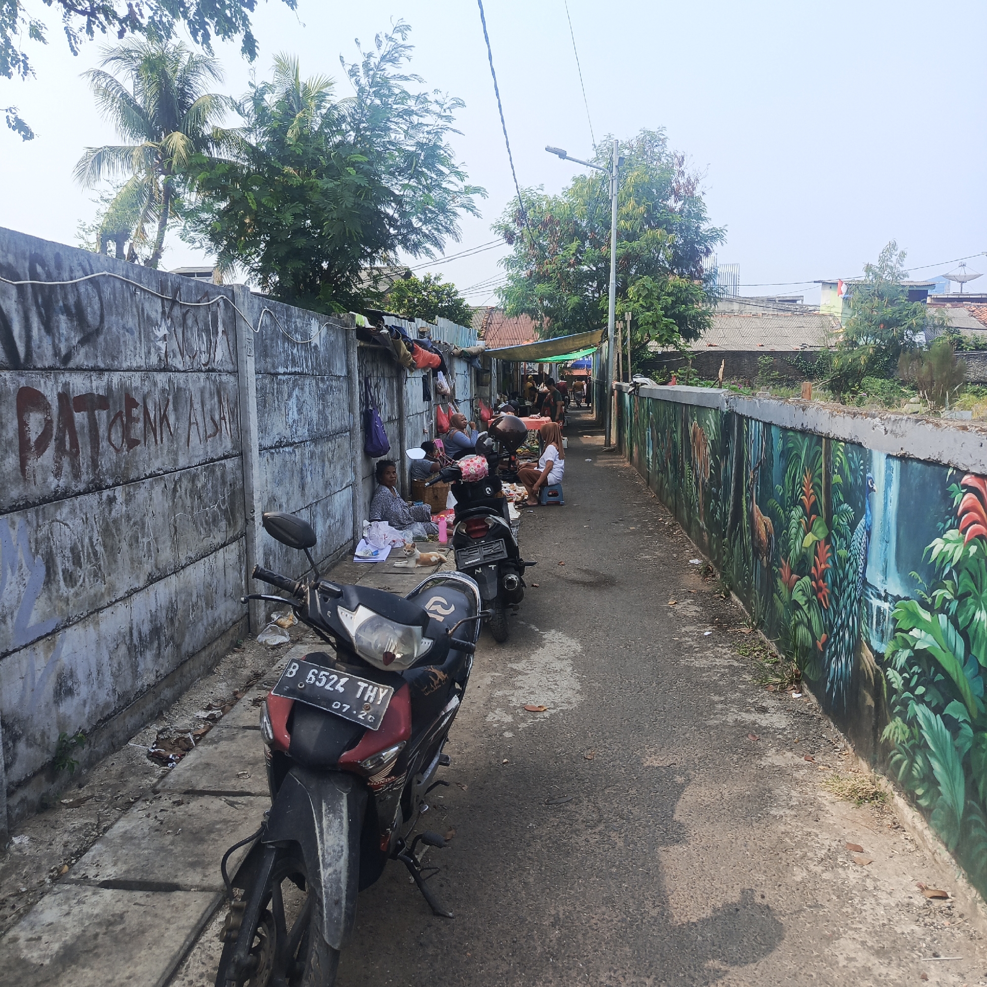 The image depicts a narrow alleyway in South Jakarta, Indonesia, bordered by concrete walls on both sides. The wall on the left is adorned with graffiti, including the text "PATOENK ALAM". Several people are seen sitting and interacting along the left wall, with some appearing to be vendors or residents engaged in daily activities. There are tarps and makeshift shelters set up along this wall. In the foreground, a red and black motorcycle is parked, its license plate partially visible as "B 6524 THY 07.20". The wall on the right side of the alley features a vibrant mural depicting a jungle scene with animals like a deer and a peacock, along with a waterfall. The alleyway itself is paved with asphalt and concrete. Trees are visible in the background, with palm trees and other foliage suggesting a tropical environment. The sky is overcast or hazy. The scene conveys a sense of a bustling, lived-in urban environment with elements of both grit and artistic expression.