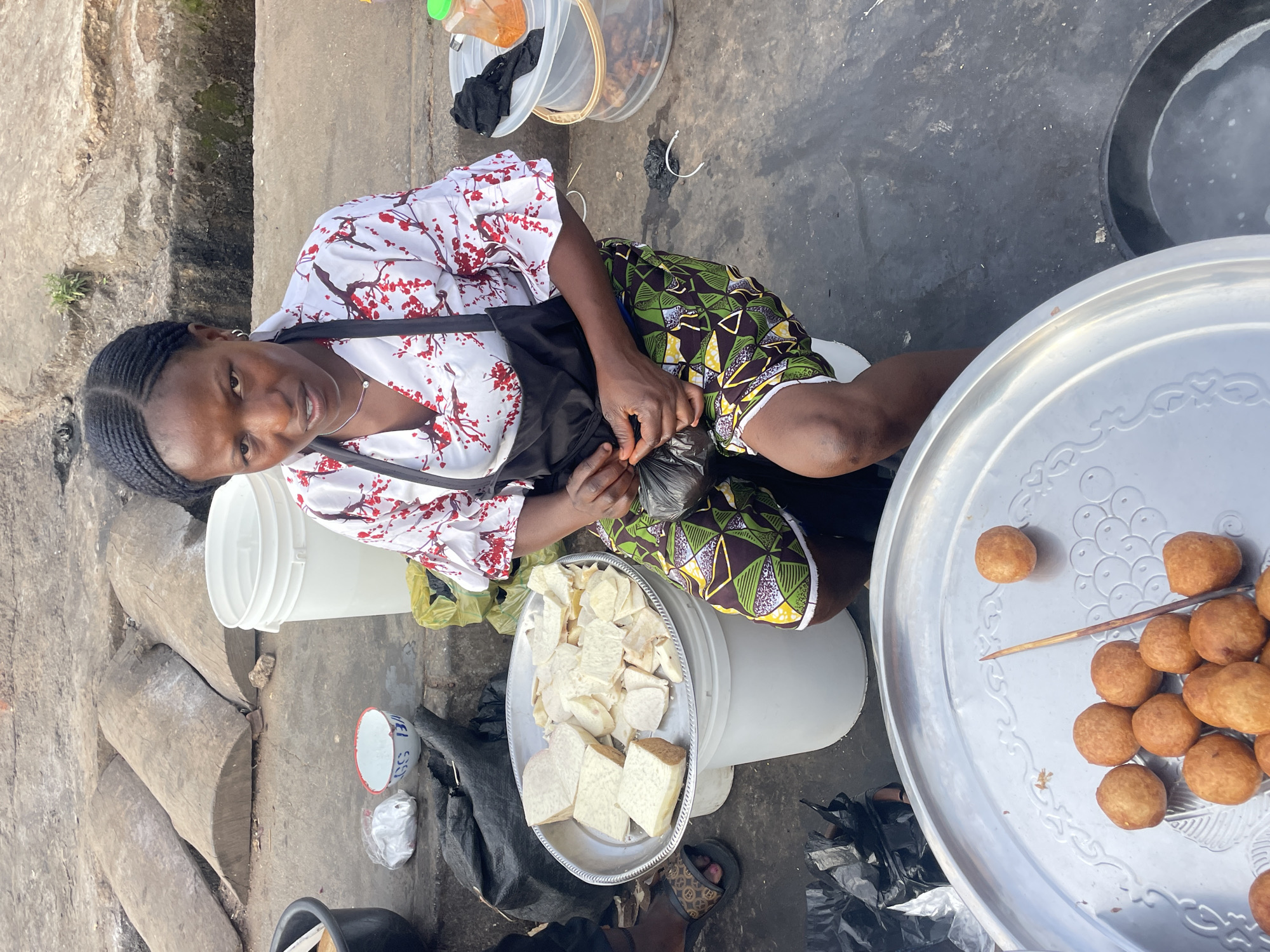 This image captures an outdoor, informal food vending scene located in Jos, Nigeria. A woman, the apparent vendor, is positioned centrally, seated on a low white plastic bucket. She has dark braided hair and is wearing a white blouse adorned with a red floral pattern and ruffled sleeves, a black apron, and patterned shorts or a skirt featuring green, yellow, black, and white geometric designs. She wears small hoop earrings and a delicate silver necklace. Her hands are occupied with a small black plastic bag.
In the immediate foreground, a large round silver tray holds numerous spherical, golden-brown fried dough items, with a slender wooden stick resting on the tray. To the woman's left, a second silver tray is laden with cut pieces of yam, presented in both larger block and smaller slice forms, resting atop another white plastic bucket. A third white plastic bucket is visible behind this setup. On the ground near the yam, a small white bowl with a red rim and a small white plastic bag are present.
To the woman's right, a transparent plastic bucket with a lid contains darker fried food items, and a small orange container with a green lid sits on its cover. The ground is an uneven concrete surface. In the background, three weathered, half-cylindrical wooden logs or concrete blocks are embedded or resting on the ground. Various black plastic bags are scattered throughout the scene.