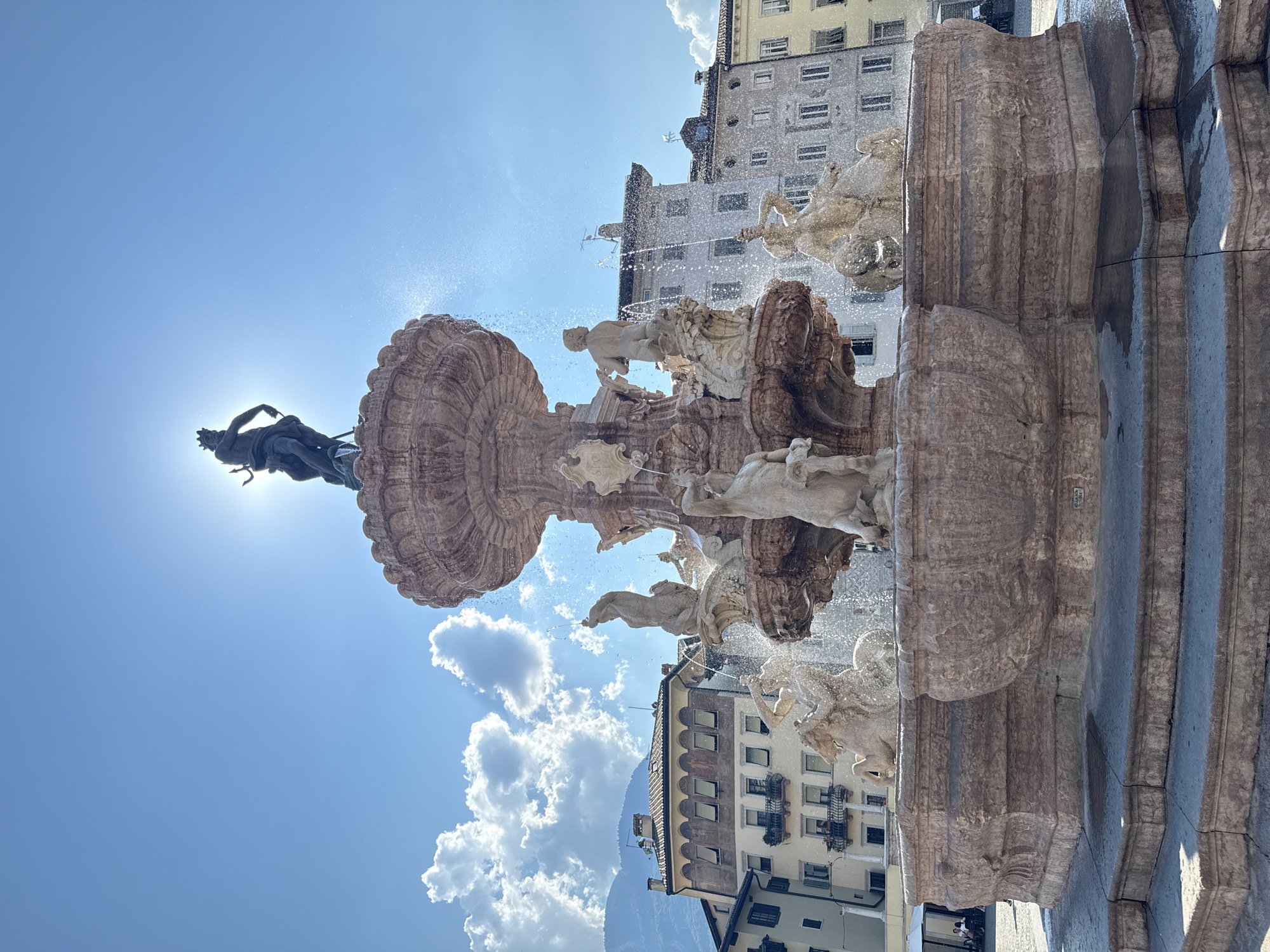 The image shows the Fontana del Nettuno (Neptune Fountain) in Piazza Duomo, Trento, Italy. The fountain is a multi-tiered structure made of pinkish-brown stone, featuring various sculptures. At the very top, silhouetted against a bright sky with a few clouds and mountains in the background, is a statue of Neptune holding a trident. Below Neptune are several sculpted figures, including what appear to be horses and human-like beings, from which water is cascading. The fountain is situated in a paved public square. In the background, there are buildings with historical architecture, some featuring balconies and awnings. The sky is bright blue with the sun partially obscured by the top of the fountain, creating a sunburst effect. A few people are visible in the far background, seated at a table under an umbrella. The overall scene depicts a sunny day in a historic Italian city square dominated by a large, ornate fountain.