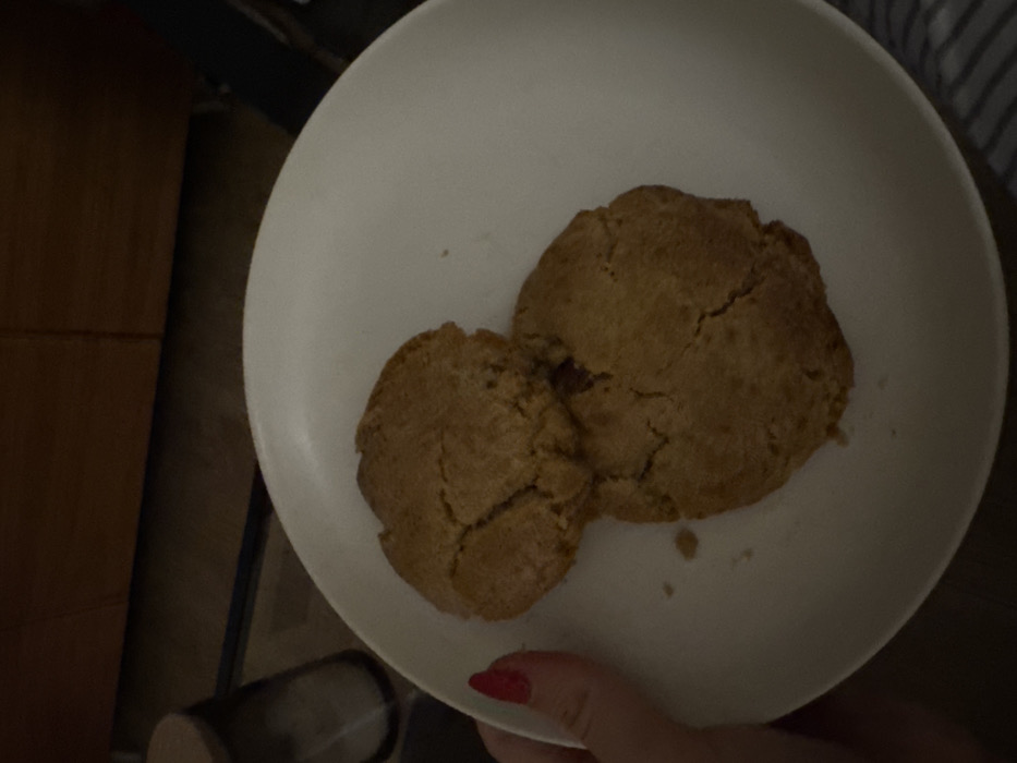 Two golden-brown cookies rest on a white plate. The cookies have a slightly cracked surface, suggesting they are baked. One cookie is slightly larger than the other. A hand with red nail polish holds the edge of the plate. The plate is positioned at an angle and the background includes a wooden surface and a striped fabric.
