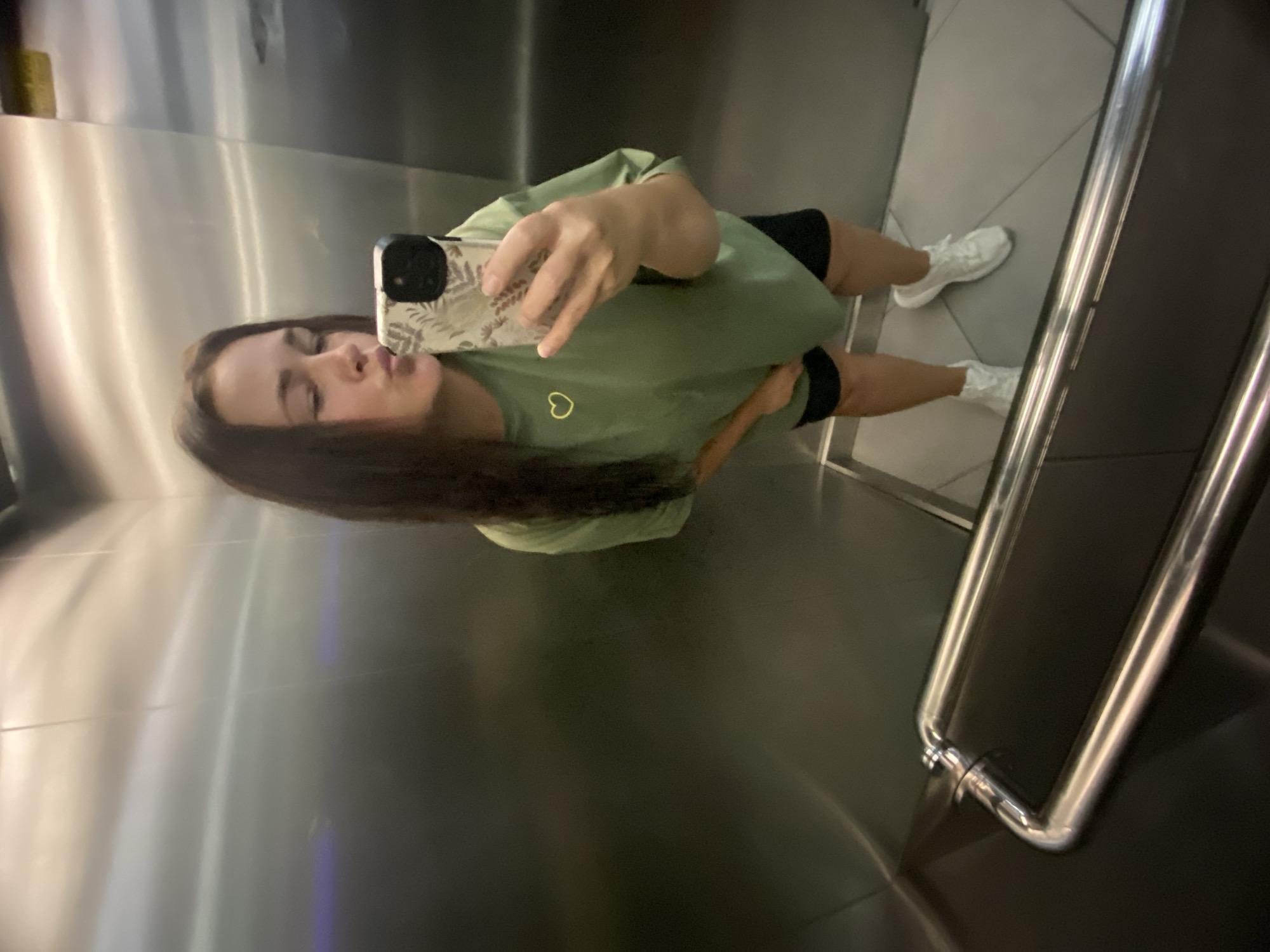 A young woman with long dark hair is taking a selfie in an elevator. She is wearing a green t-shirt with a yellow heart emblem and black shorts, along with white sneakers. Her face is turned towards the camera as she puckers her lips for a kiss. The elevator walls are made of polished metal, reflecting the scene. A silver handrail is visible in the foreground. The floor of the elevator is tiled in a light grey color. The image appears to be taken in Sochi, Russia.