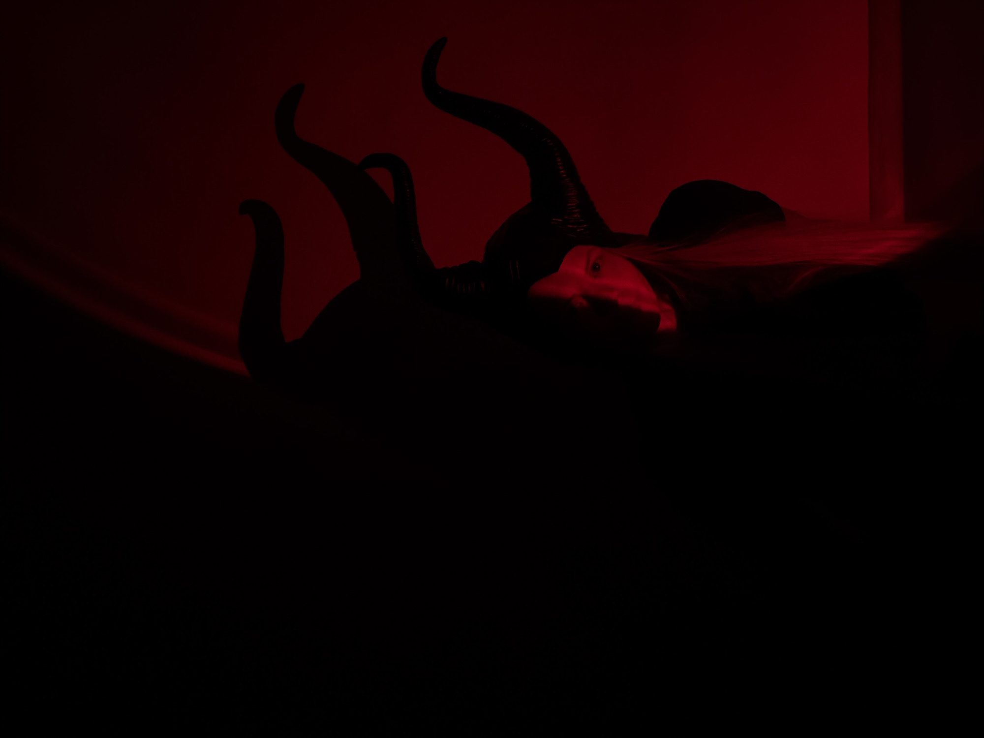 The image displays a dimly lit, dramatic scene dominated by deep reds and blacks. A human figure is positioned lying down, with most of their body obscured by shadow. Only a portion of their face – an eye, nose, and mouth area – is faintly illuminated by a strong red light, revealing pale skin and dark hair that catches the red hue.
Emanating from around the figure's head are several prominent, dark, curved forms resembling large horns or tentacles. These objects are in sharp silhouette, but closer inspection reveals some texture, suggesting they are part of an elaborate headpiece or costume. The background is a solid red color, possibly a wall or draped fabric, with subtle light variations hinting at folds or light sources. On the far right, a faint vertical structure, possibly a pillar or architectural element, can be observed. The foreground is entirely consumed by shadow, contributing to the dark and mysterious ambiance.
The setting appears to be indoors, likely a studio or controlled environment, given the artificial lighting. The overall mood is intense, ominous, and theatrical. No specific activity beyond posing or resting is discernible. There is no visible text within the image. The image originates from Izhevsk, Russia.