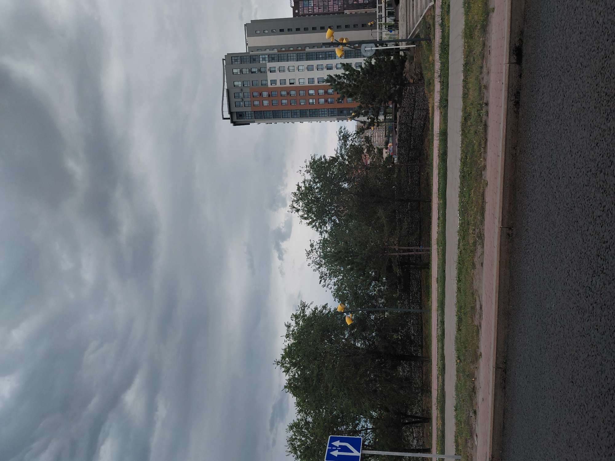 A street scene in Qarağandı city administration, Kazakhstan under a cloudy sky. In the foreground, there is a paved road and a sidewalk. Beyond the sidewalk is a strip of grass and then a row of trees. Behind the trees, there are several modern multi-story buildings. A blue traffic sign with two arrows indicating lanes merging is visible on the left side of the image. Several yellow streetlights are positioned along the roadside. The overall impression is that of an urban environment on an overcast day.