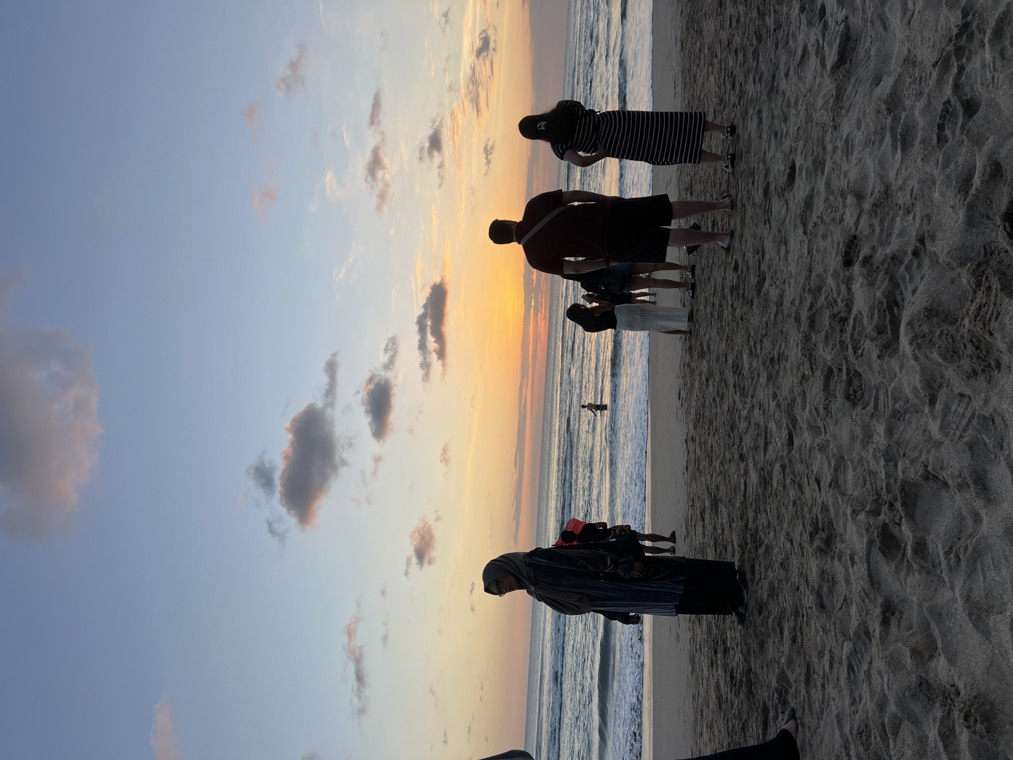 A group of people are gathered on a sandy beach in Kuta, Indonesia, watching the sunset. The sky is a gradient of blue and orange, with scattered clouds. The ocean is calm, with gentle waves rolling onto the shore. Several individuals are visible in the foreground and mid-ground, facing the ocean. One person in the foreground, wearing a dark headscarf and a long, dark garment, stands facing left. To their right, a group of three people are also looking towards the sunset. Further back, near the water's edge, more individuals are present. The sand is light brown and textured, with footprints visible. The overall scene depicts a peaceful evening at the beach.