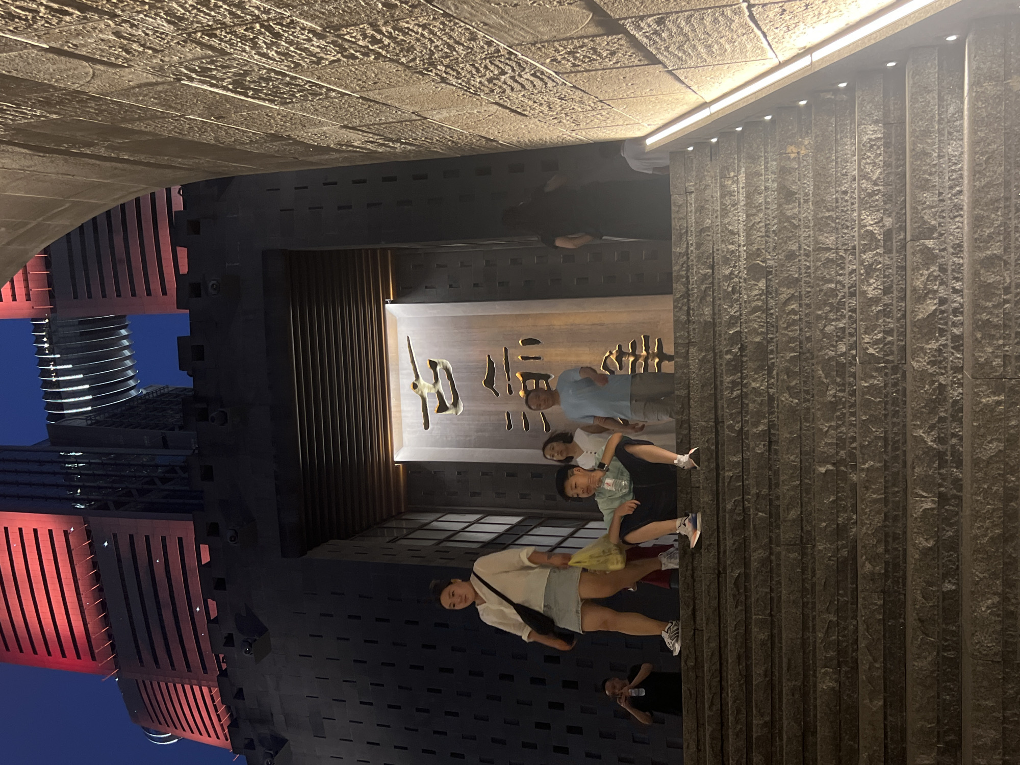 A group of people are on a stone staircase leading up to a large, dark entryway. The entryway is framed by a stone structure that resembles ancient architecture, with the modern Nan'an District's illuminated buildings visible in the background. A woman in a white shirt and denim skirt, carrying a yellow bag, walks up the stairs. A young boy in a green shirt sits on the stairs, drinking from a water bottle. Another woman and a man are also on the stairs, heading up. A man in a black shirt stands on a lower step, holding a phone. The scene is lit by artificial lights, highlighting the textured stone of the staircase and walls, and the glowing red of the buildings.
