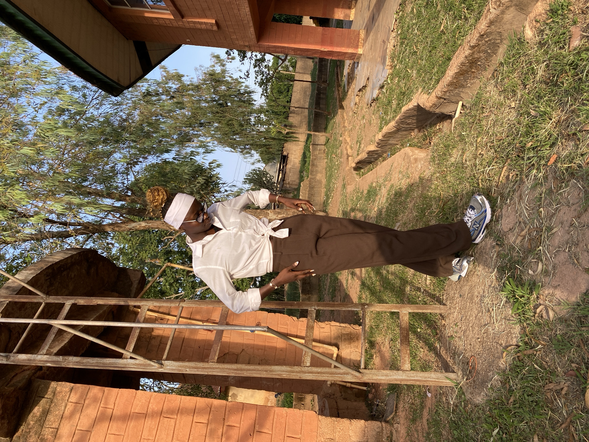 The image depicts a person standing outdoors in Vom, Nigeria. The person is wearing a white shirt tied at the waist, brown trousers, and athletic shoes. They also have a white head covering and glasses. They are standing near a brick structure with scaffolding, and in the background are trees and other buildings. The ground is a mix of grass and dirt.