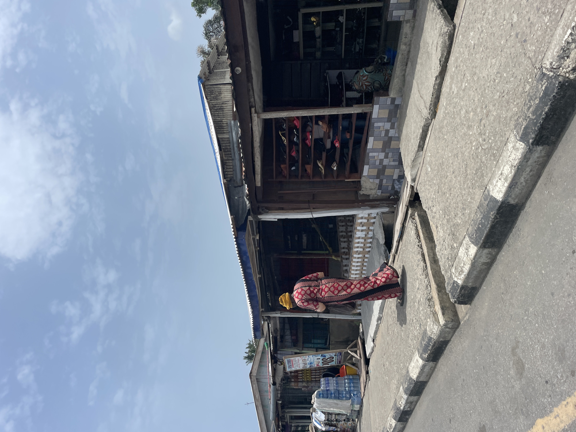 A street scene in Badagry, Nigeria shows a woman in a red patterned robe and yellow headscarf standing on the sidewalk next to a storefront. The storefront has a corrugated metal roof, wooden frame, and displays shoes on shelves. Beside the woman, bottles of water are stacked. The sky is blue with scattered clouds. The road is asphalt with a painted curb.