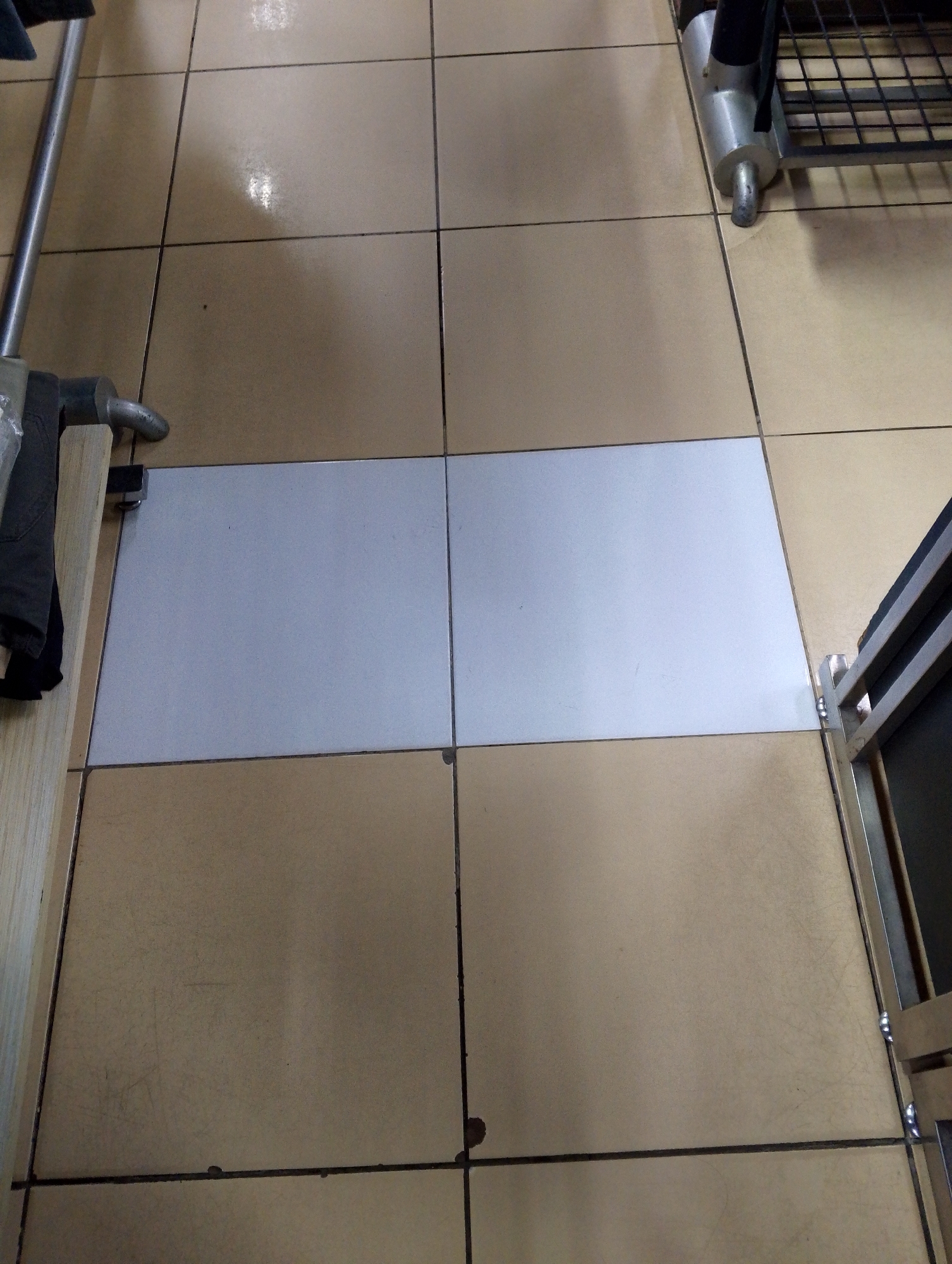 The image depicts a section of tiled flooring in what appears to be a commercial or public space, possibly a store or a hallway, in Batam, Indonesia. The tiles are predominantly a light beige or tan color, laid in a grid pattern. A contrasting strip of lighter, possibly white or pale blue, tiles is set into the floor, running horizontally across the frame. To the left, a metallic pole, likely part of a display rack or structure, is visible, along with a piece of dark fabric. To the right, a portion of a metal shelf unit with a black wire grate is present. The lighting suggests an indoor environment, potentially with overhead illumination causing some reflections on the glossy tile surface. There are no individuals visible in the image. The scene is clean and orderly, with no obvious signs of damage or disarray. The overall impression is that of a utilitarian and functional area within a building.