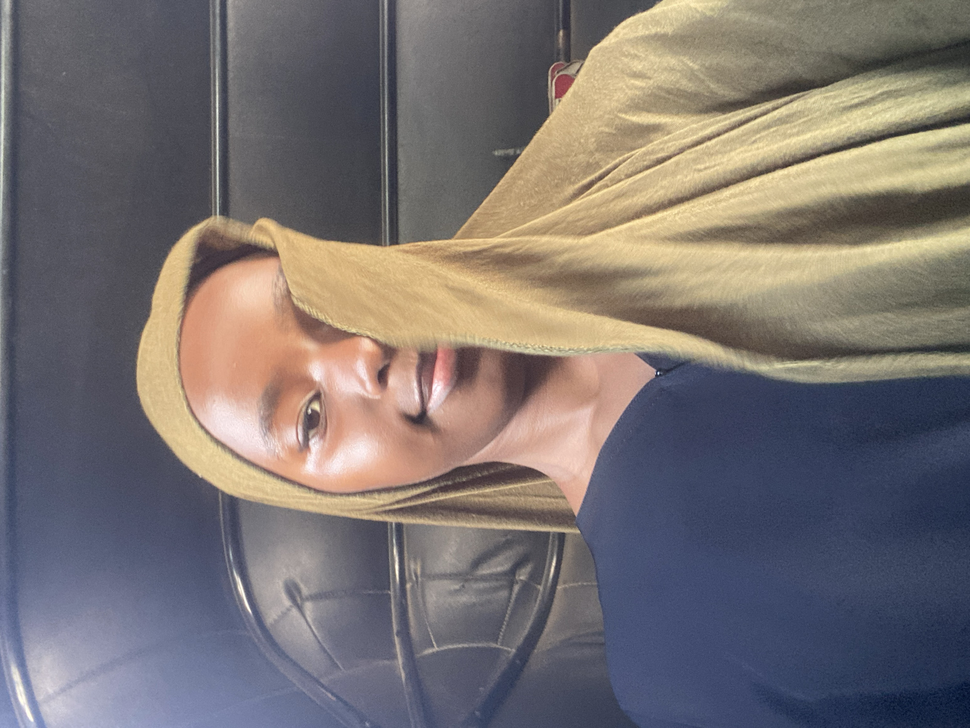 A person wearing a gold-colored headscarf and a navy blue shirt is seated in what appears to be a bus or vehicle interior. The seat behind the person has a black leather or leatherette upholstery. The person is facing forward, partially obscured by the headscarf. The location and its relationship to Dar es-Salaam, Tanzania, cannot be determined from the information available in the image.