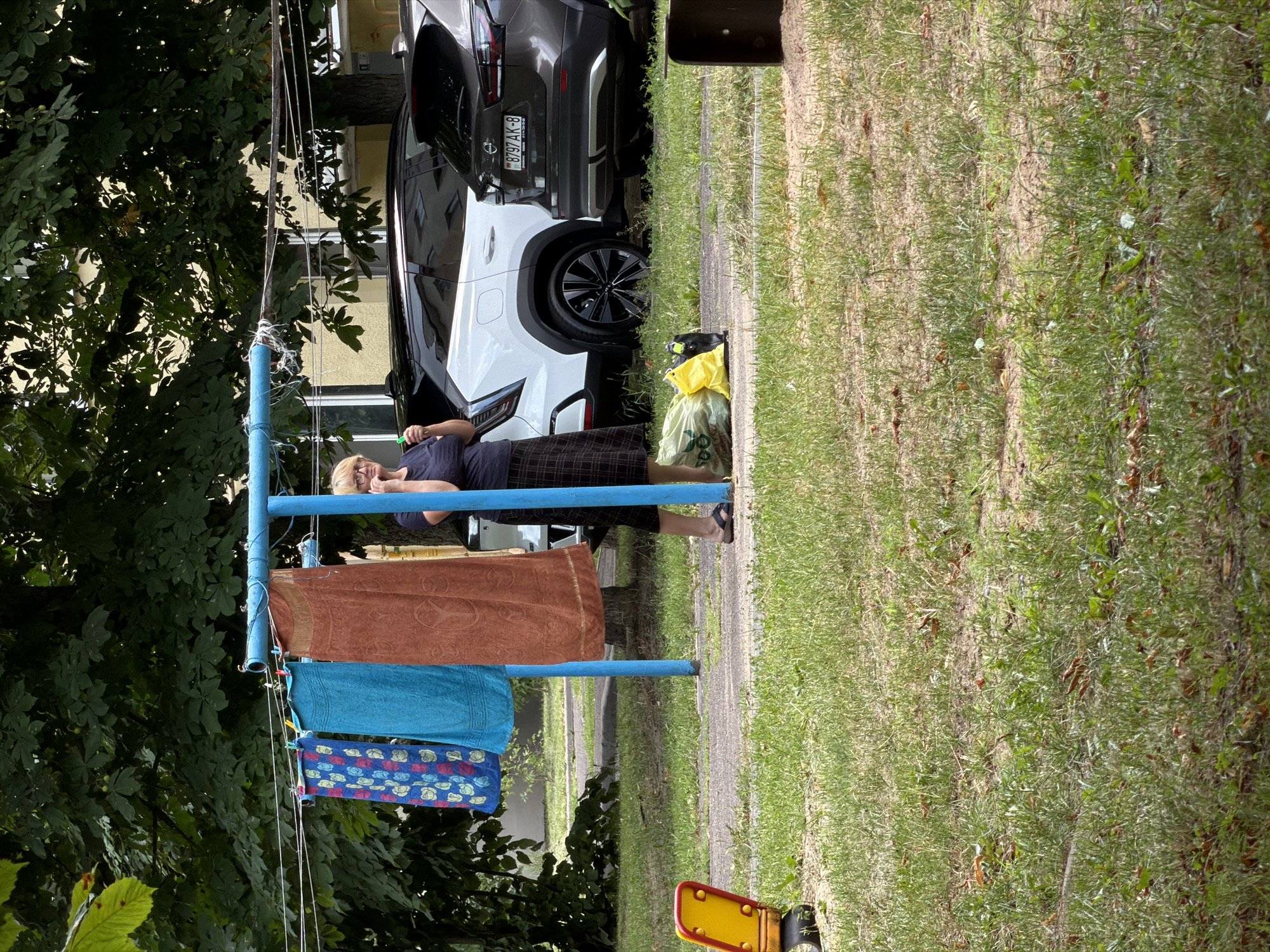 An outdoor, eye-level, medium shot shows a woman standing in front of a clothesline with laundry hanging on it, near parked cars in what appears to be a residential courtyard in Minsk, Belarus. The woman, with blonde hair and glasses, is wearing a dark blue shirt and a plaid skirt and is holding a cell phone to her ear. Three towels hang on the blue clothesline: a blue towel with a floral pattern, a solid turquoise towel, and a rusty brown towel with a circular pattern. Behind the clothesline and to the right of the woman, a white SUV and a dark grey SUV are parked. The ground is covered in grass and a gravel path. In the foreground, a portion of a yellow and red children's toy is visible on the left. The background features lush green trees and the yellow facade of a building. The overall scene suggests a typical summer day in a residential area.