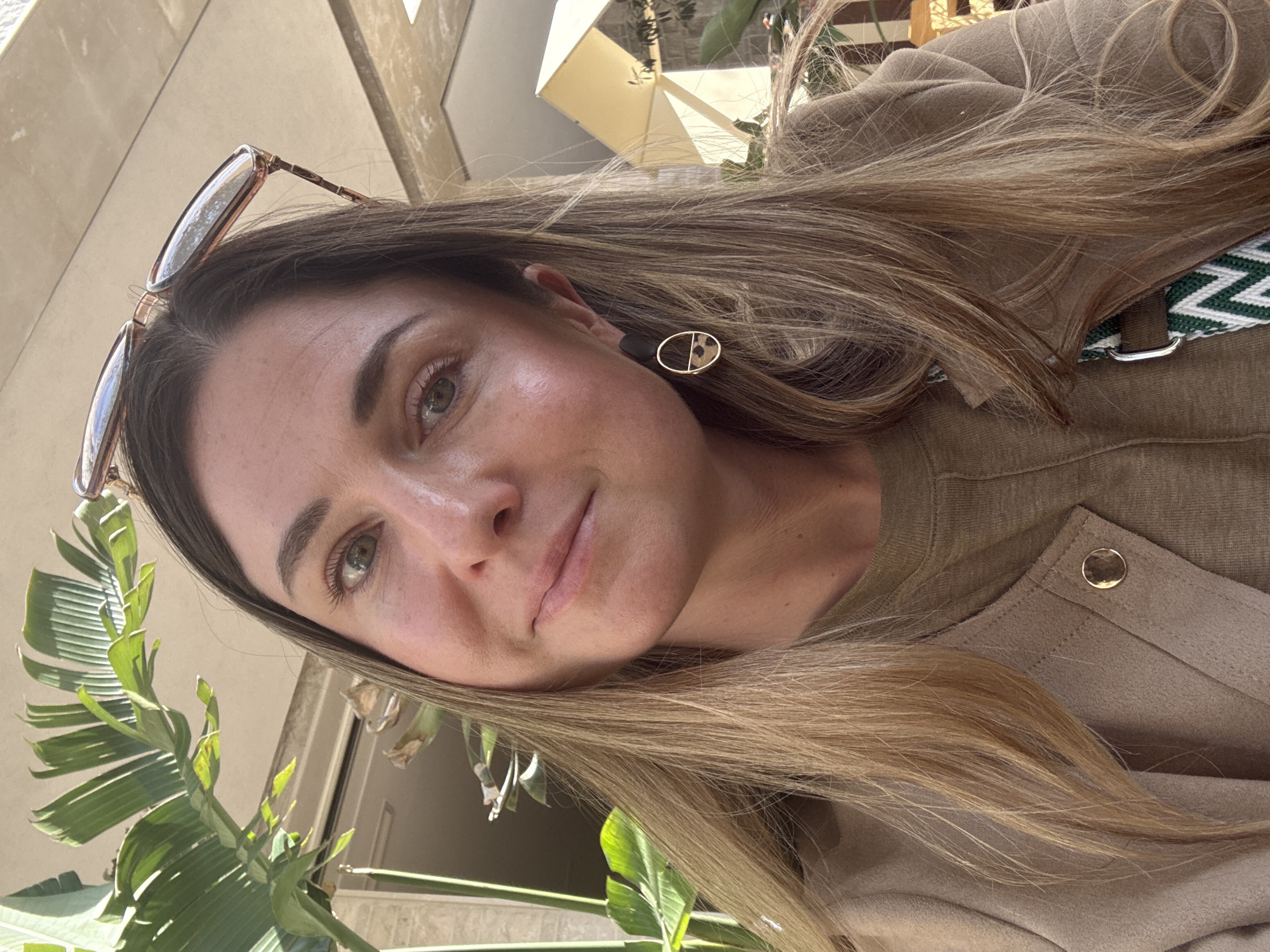 The image depicts a woman in Đenovići, Montenegro, posing outdoors on a sunny day. She has light brown hair, fair skin, and green eyes. Sunglasses are perched on her head. She is wearing a brown top with a decorative button and geometric earrings. Part of a green and white strap is visible over her shoulder. In the foreground, lush green plants fill the bottom left corner, and in the background, a glimpse of building facades suggests an urban environment.