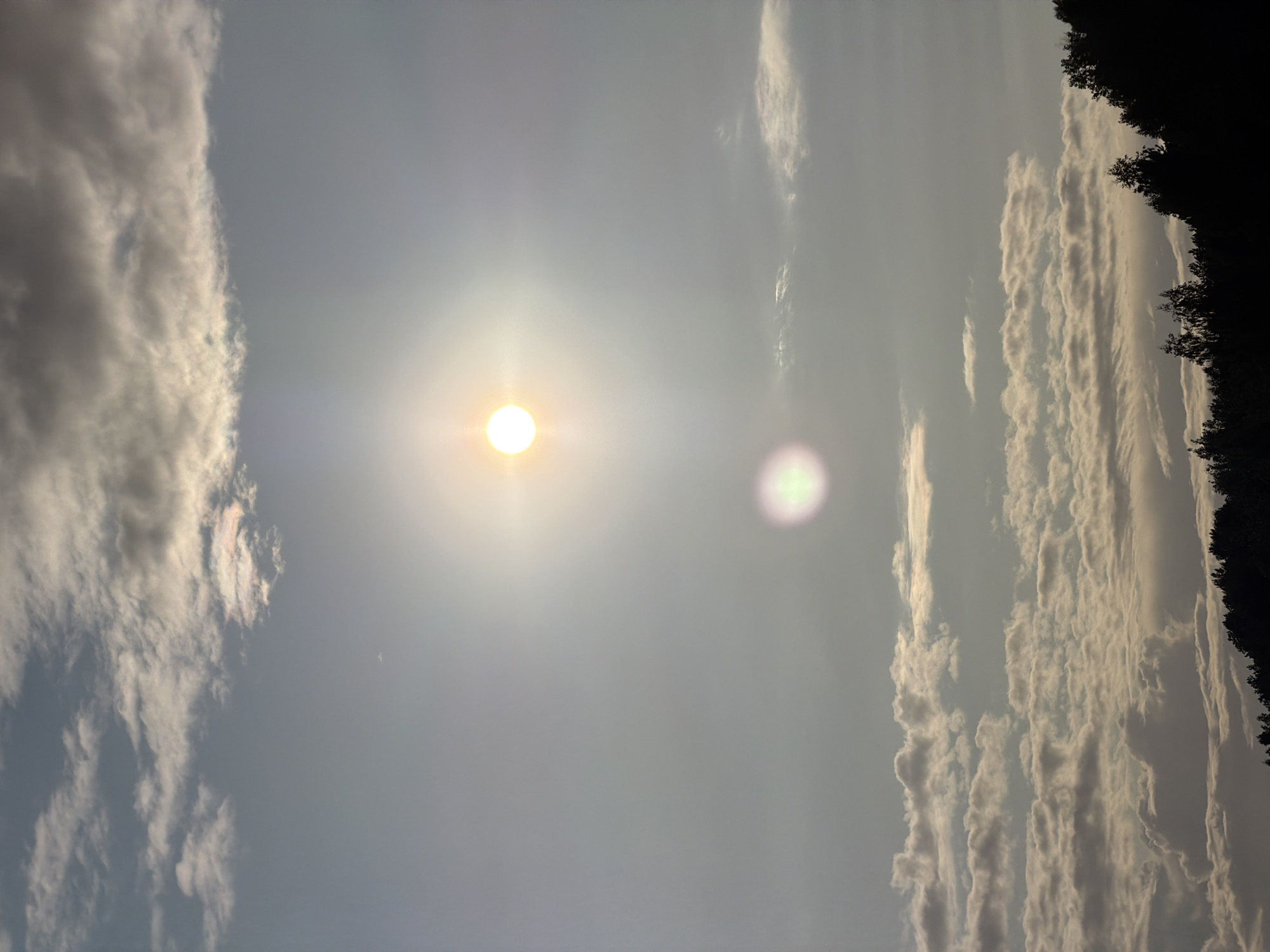 The image depicts a sky with scattered clouds and the sun prominently featured. The sun is a bright, glowing orb, casting a soft light and creating a lens flare. Below the sun, a distinct, smaller orb of light, possibly another lens flare or atmospheric phenomenon, is visible. The clouds are varied in texture and density, with some appearing wispy and others more solid. Towards the bottom of the frame, a dark silhouette of trees and possibly a hillside suggests a landscape feature. The overall impression is that of an outdoor scene, likely taken during daytime. Without further context, specific details about Budachi, Belarus, cannot be determined from the visual information alone.