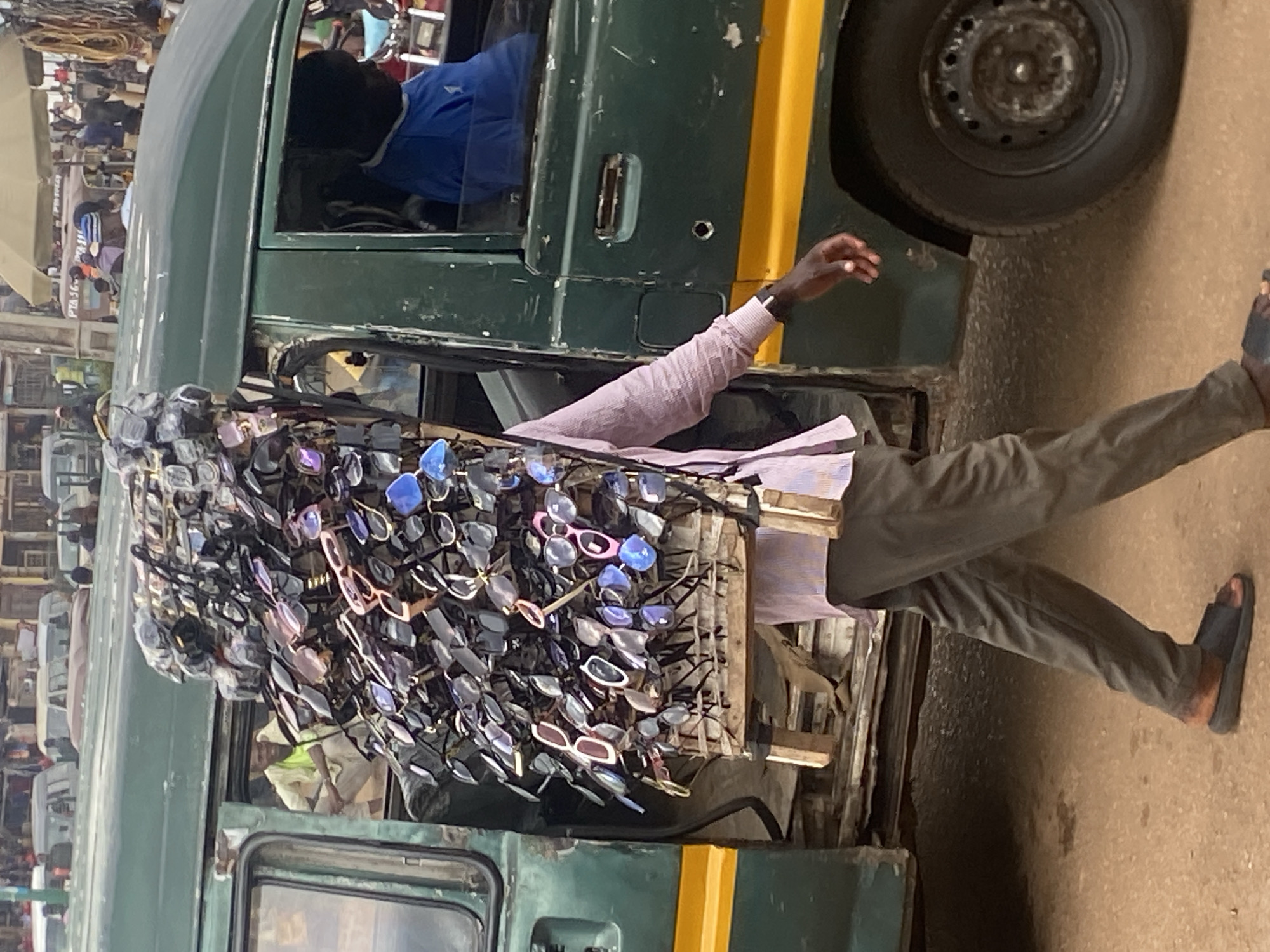 A man walks with a large display of sunglasses attached to a wooden rack, which he carries on his back. He is wearing a light pink shirt and dark pants, and black sandals. He is walking alongside a green van with a yellow stripe. The van's door is open, and a person can be seen sitting inside. The scene appears to be a street in Bukuru, Nigeria, with other vehicles and people in the background, suggesting a busy marketplace or roadside. The ground is a dusty, reddish-brown color.