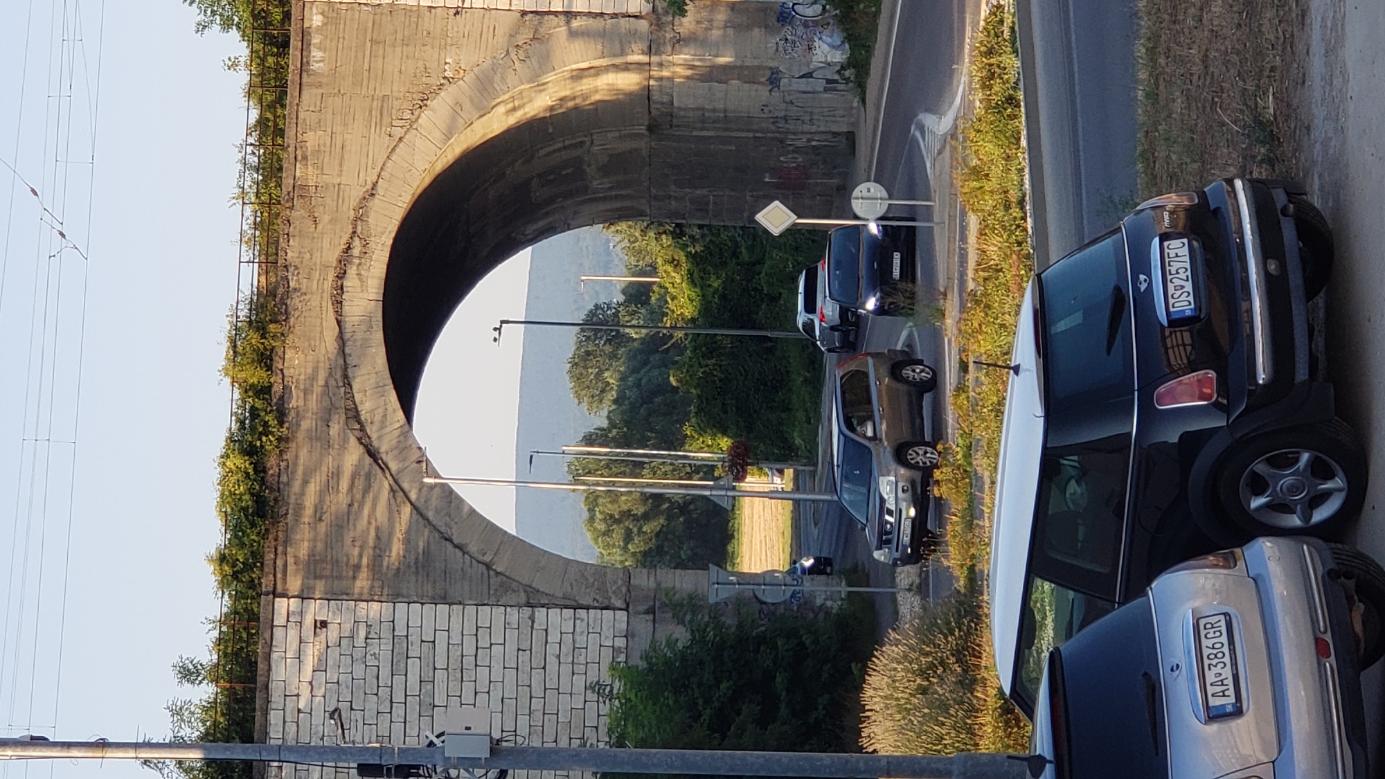 The image shows a view through an arch of a stone viaduct, likely for a railway, in Bratislava, Slovakia. The viaduct is constructed of light-colored stone blocks and concrete, with vegetation growing along its top edge. Through the arch, a road is visible with several cars driving. In the foreground, two Mini Cooper cars are parked, one silver and one black. The silver Mini has the license plate "AA 386 GR". The black Mini has the license plate "DS 257FC". The road has streetlights and traffic signs. The background shows trees, hills, and a clear sky. The lighting suggests it is either early morning or late afternoon. The overall impression is an urban landscape with a mix of historic infrastructure and modern vehicles.