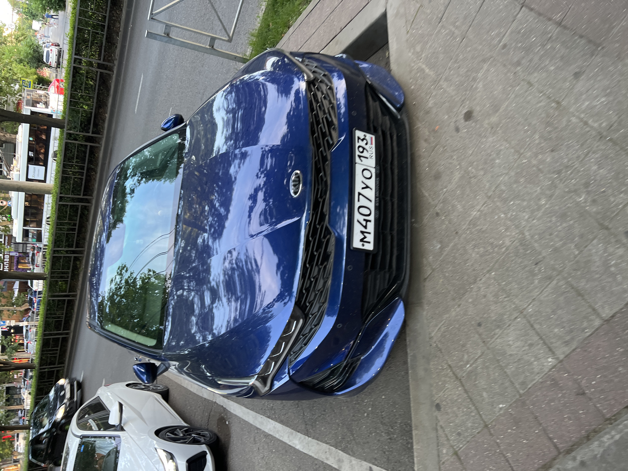 A dark blue Kia K5 is parked in a marked parking space on a street in Krasnodar, Russia. The car is the primary subject, facing the camera, with its headlights, grille, and license plate clearly visible. The license plate reads "M407YO 193 RUS". To the left of the Kia, a white sedan is also parked. The street is paved with asphalt, and there is a sidewalk made of interlocking pavers in the foreground. In the background, a hedgerow runs along the street, and beyond that, there are trees and buildings, including a bus with the number "271" displayed. There are a few people walking in the distance. The lighting suggests it is daytime, possibly late afternoon, with shadows cast by the trees.