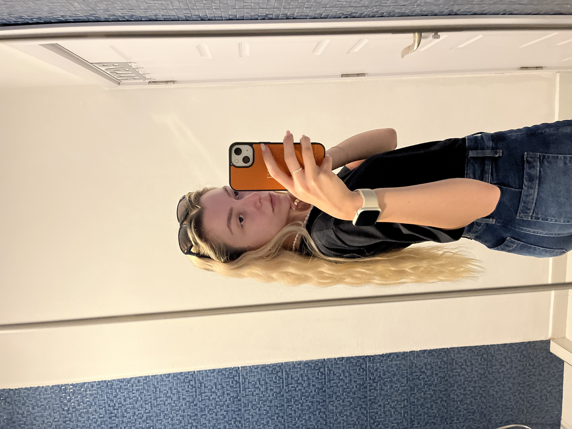 A young person is captured taking a mirror selfie in an indoor setting. The individual, appearing to be a young woman with long, wavy blonde hair, is looking directly into the smartphone she holds with her right hand. The phone has a distinctive orange case with two visible camera lenses. She is wearing a black t-shirt, blue denim jeans, and a white smartwatch on her left wrist. Sunglasses are pushed up onto her head, and a light-colored beaded necklace is visible around her neck. A delicate ring is worn on her right ring finger.
The reflected background behind the person shows a predominantly white wall or surface. Below this white area, there is a band of blue mosaic tiles, suggesting the location might be a bathroom or a similar tiled space. A white door, complete with visible hinges and a silver handle, is reflected in the upper portion of the mirror, indicating an adjacent room or cabinet. The lighting appears bright and even, consistent with indoor illumination. The overall image is a casual self-portrait taken in what appears to be a well-lit personal space. No discernible text is visible in the image.