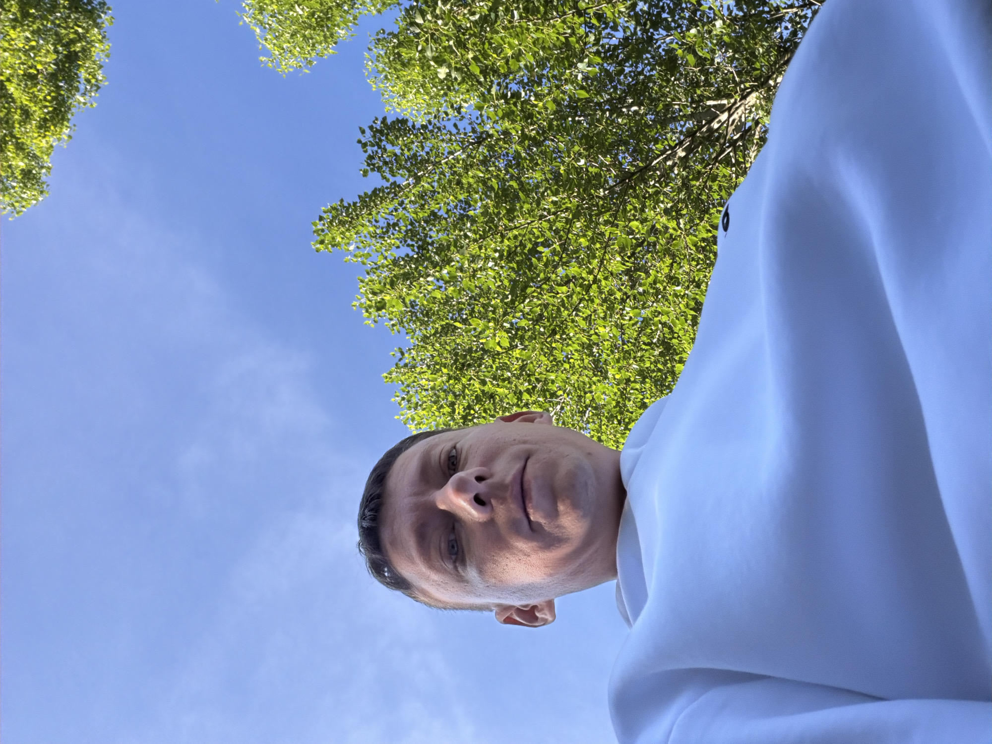 A low-angle, close-up shot shows a man wearing a white hoodie, looking up towards the sky. His face is partially obscured by the perspective. Green foliage from trees is visible against a clear blue sky. The image is shot outdoors, possibly during the daytime, with natural lighting. There are no discernible Moscow landmarks or specific environmental details that would indicate its location within the city.