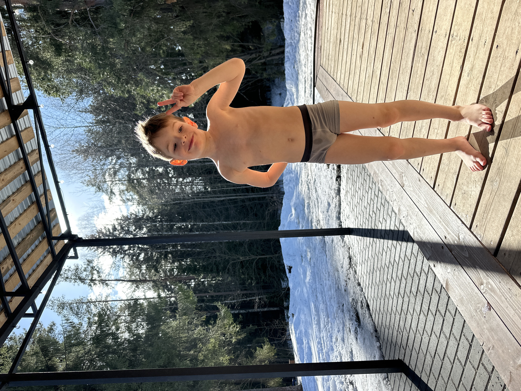 The image captures a playful young boy standing outdoors on what appears to be a sunny but cold day, given the presence of snow. The photo is taken from a slightly low angle, looking up at the boy and the sky.
The main subject is a fair-skinned boy with light brown hair, appearing to be in elementary school age. He is shirtless and wearing only grey boxer briefs with a black waistband. A noticeable wet spot is visible on the front of his underwear. He is smiling brightly at the camera and making a peace sign with his right hand, while his left hand rests on his hip. His face is illuminated by bright sunlight, creating a halo effect around his hair. He is barefoot, standing on a wooden deck.
The setting is an outdoor area featuring a wooden deck with horizontal planks, which occupies the right side of the frame. To the left of the deck, there's a grey paved area, likely interlocking pavers or bricks, partially covered in snow. Beyond this, a significant amount of white snow covers the ground, leading into a dense forest of tall trees, many of which appear bare, suggesting winter or early spring. Evergreen trees are also visible.
In the upper left portion of the image, part of a structural element is visible. It features dark metal framing and wooden slats, possibly part of a roof, pergola, or shelter. String lights are strung along one of the beams. The background showcases a clear, bright blue sky with sparse clouds, indicating good weather despite the snow.
The lighting is bright and sunny, casting distinct, long shadows from the structures and the boy onto the deck and paved area, suggesting either morning or late afternoon. The overall mood is cheerful and active, with the boy striking a joyful pose despite the cold environment. There is no visible text in the image.