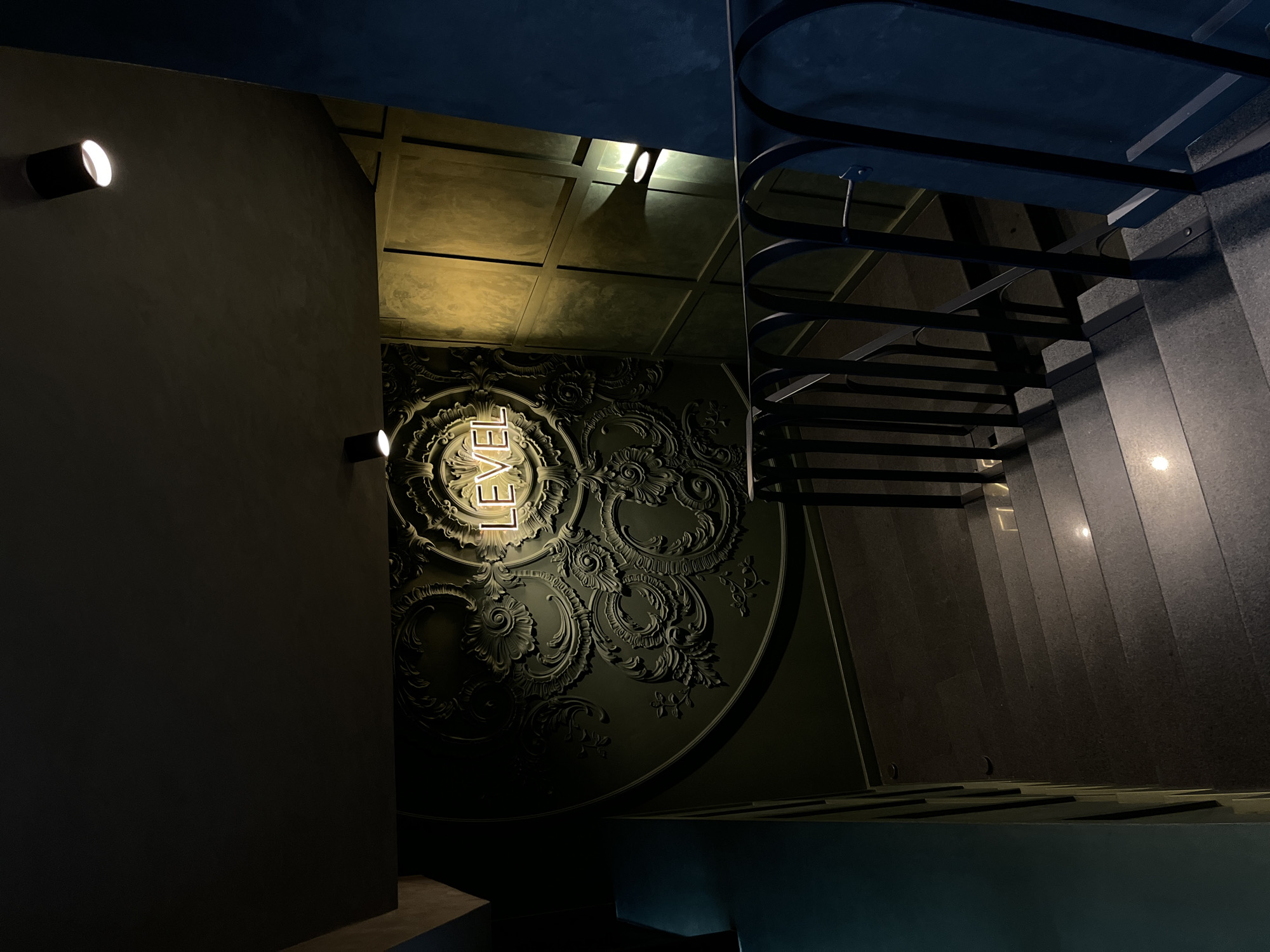 The image shows an interior view of a building with a staircase. The interior has a dark color palette, with accents of light highlighting features. On the left, a wall with two spotlights is visible. Adjacent to it is a wall with an ornate, circular design featuring the word "LEVEL" vertically in illuminated letters. To the right, a staircase with a black metal railing extends upwards. The location is Sochi, Russia.