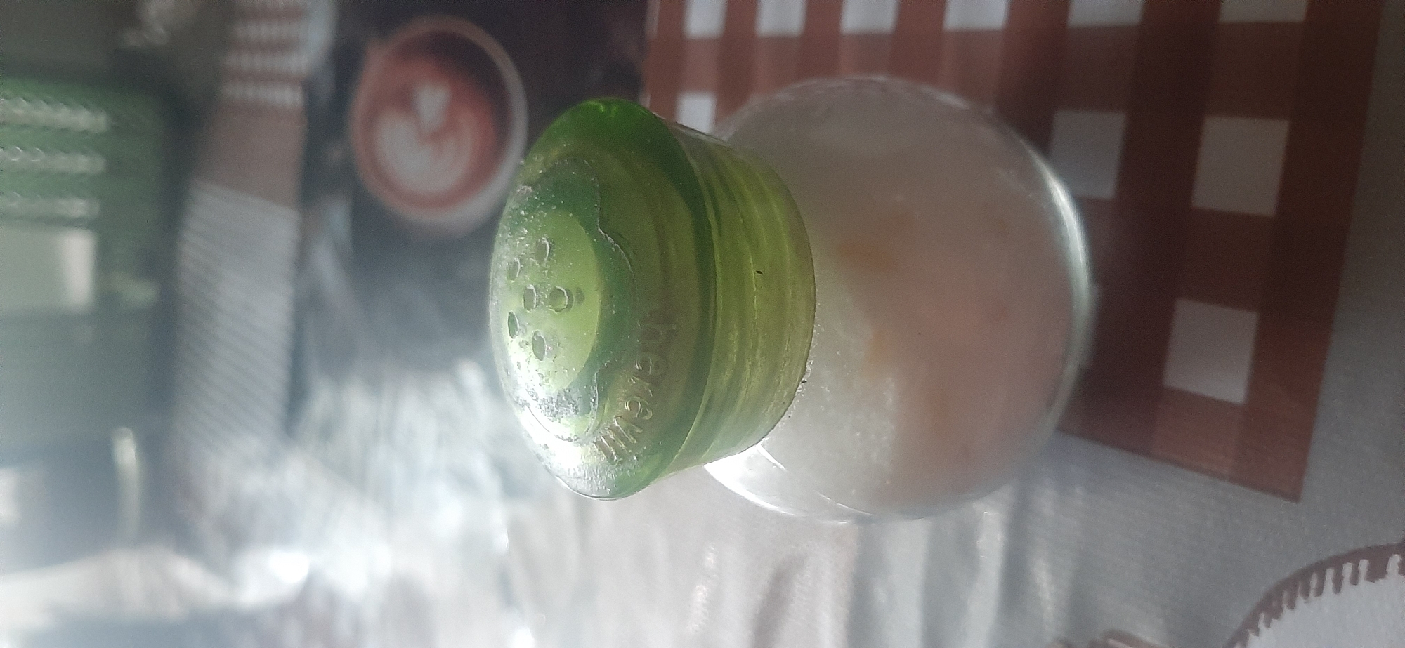 A close-up shot shows a clear glass salt shaker with a green plastic lid containing multiple holes, suggesting it is for dispensing salt. The shaker is partially filled with a fine, granular substance that appears to be salt, possibly with a slightly pinkish hue. The salt shaker sits on a surface covered by a patterned tablecloth, which has a checkered design with brown and white squares. In the background, out of focus, a portion of a coffee cup with latte art is visible on a dark surface, and a blurred green object, possibly a glass or container, is also present. The lighting suggests an indoor setting. The image does not contain any information about Yessentukskaya, Russia.