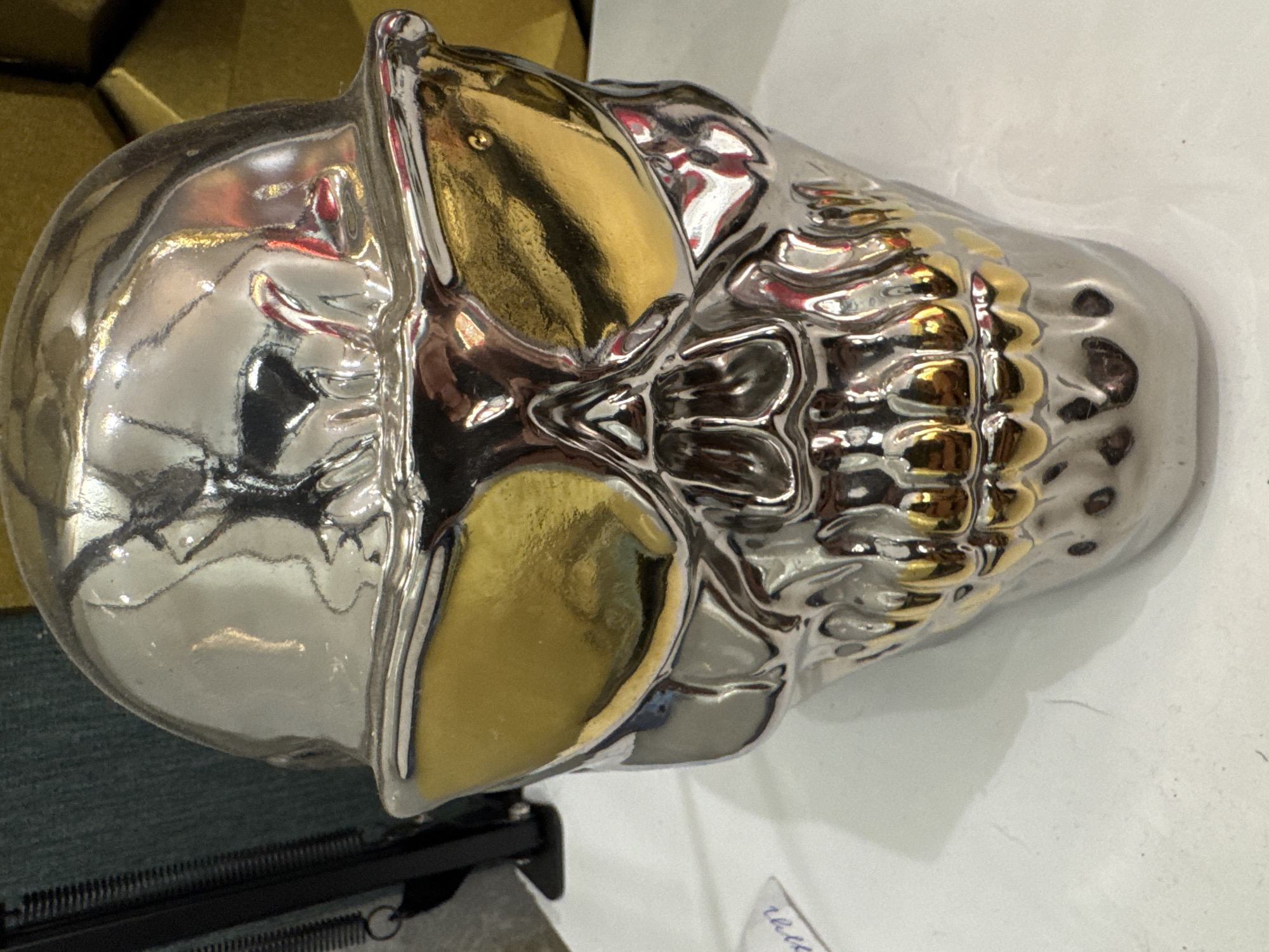 A silver and gold skull-shaped ornament sits on a white surface. The ornament has a reflective, polished finish with gold accents on the eye sockets and teeth. The background is blurred, showing hints of dark fabric and yellow material. There are no people or actions depicted in the image. The image does not contain any information about the city of Protasovo, Russia.