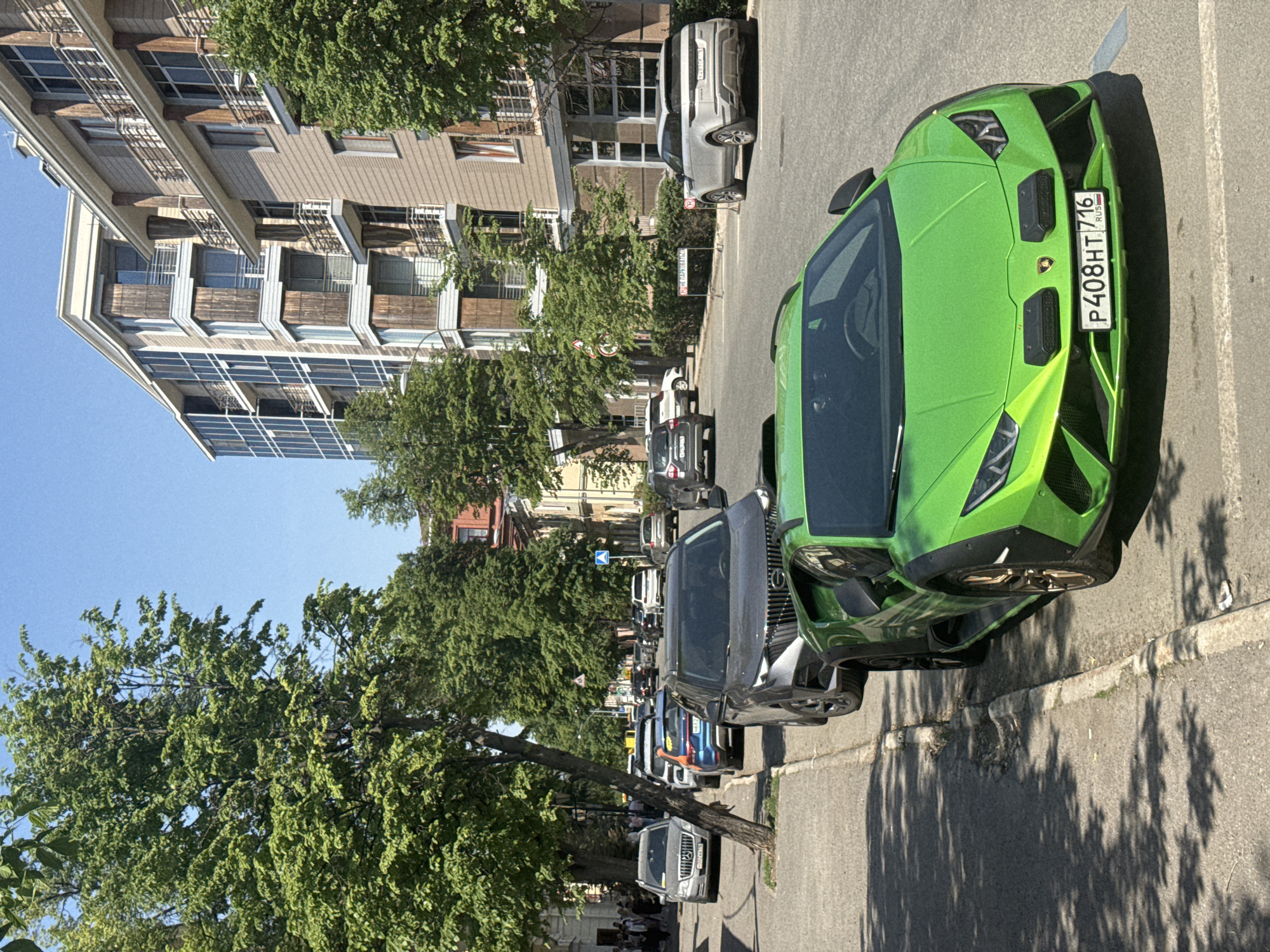 The image shows a bright green Lamborghini Huracan parked on a street in Kazan, Russia, as identified by the license plate. The license plate reads "P408HT|116" with a "RUS" marking. The car is parked along a curb on a paved street with other parked cars visible further down the street. Trees line the street, casting shadows on the ground. A multi-story building is visible in the background on the left side of the frame. The street appears to be a residential or commercial area. The sky is blue and the image is taken during daylight hours.