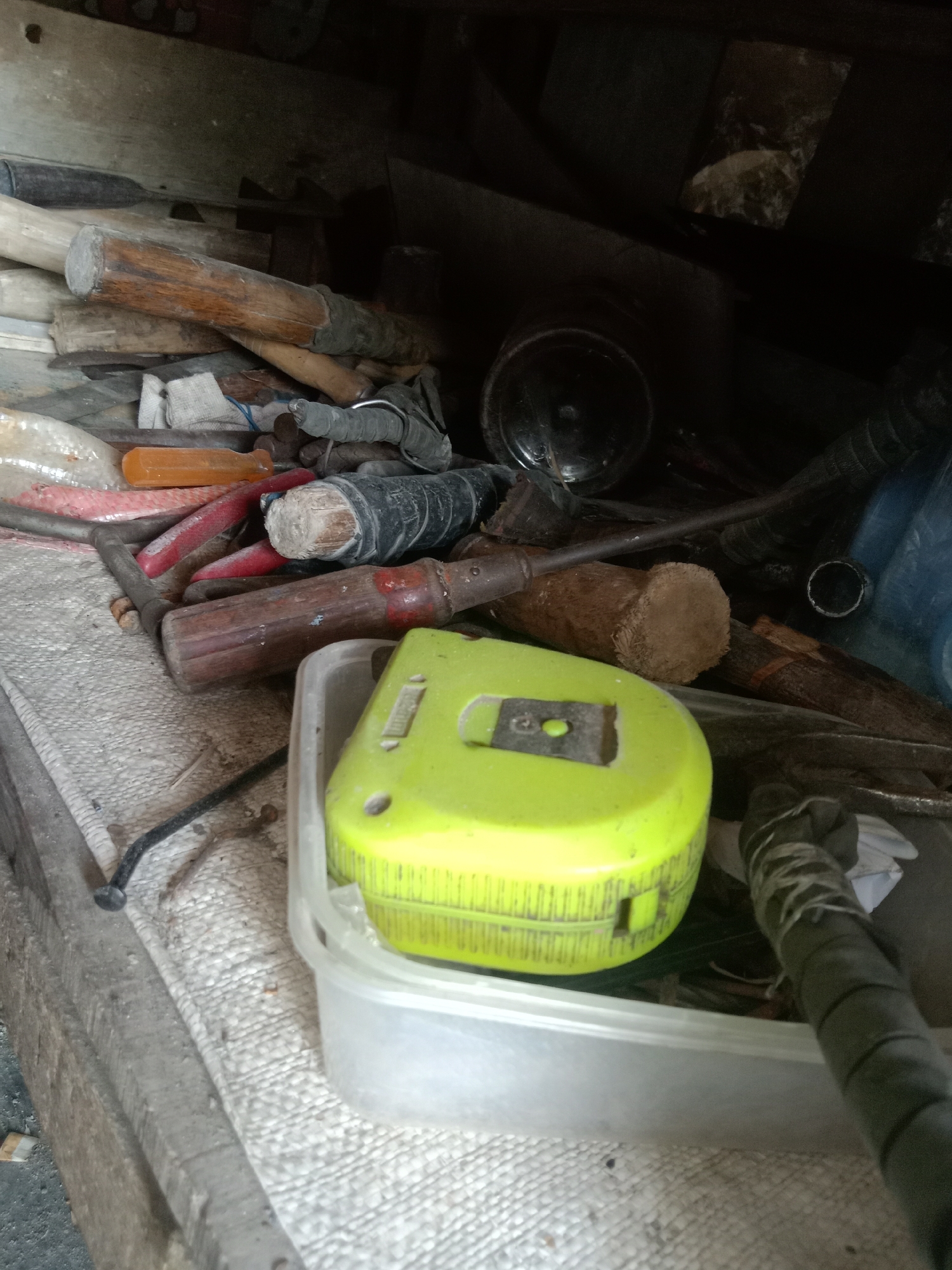 The image depicts a cluttered collection of tools and materials, possibly in a workshop or storage area. The foreground is dominated by a bright yellow tape measure resting inside a clear plastic container. To the left of the tape measure, a textured, light-colored surface, likely a mat or cloth, covers a wooden surface. Various tools are visible throughout the scene, including what appear to be screwdrivers with red and orange handles, a wooden-handled hammer or chisel, and other metal tools. Pieces of wood and what looks like a dark-colored bottle or container are also present. The background is dark and indistinct, suggesting a dimly lit environment. The overall impression is one of disorganization and the presence of work-related items. There is no indication of a specific location within Stiaji, Indonesia, or any people in the image.