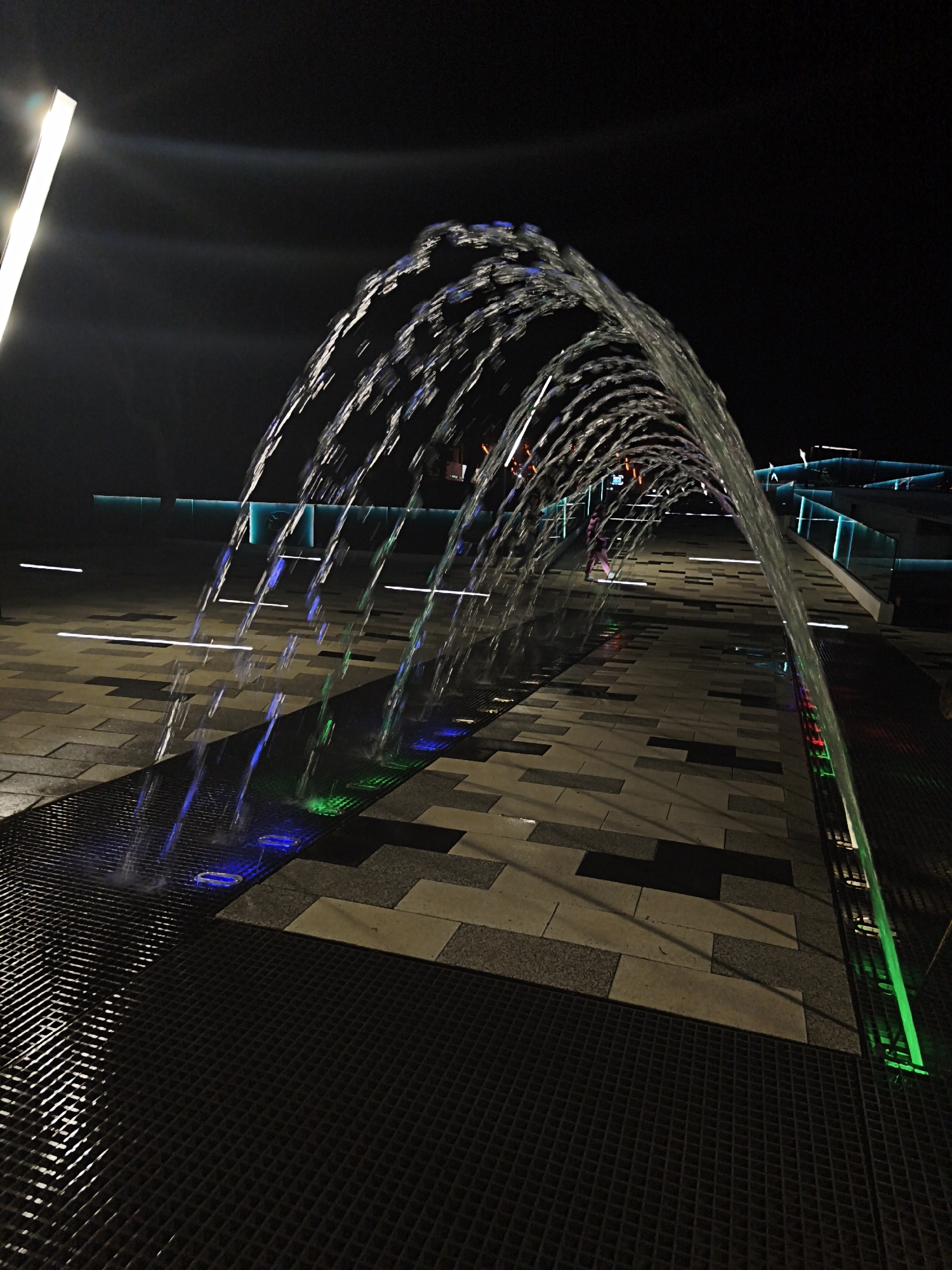 The image depicts a water fountain in Myskhako, Russia, at night. The fountain is comprised of rows of water streams arching over a walkway. The walkway is paved with light and dark colored rectangular stones. Beneath the water jets are grates and platforms illuminated by blue and green lights. A tall light pole is visible on the left. A person in pink clothing is walking through the fountain in the distance. There are illuminated teal structures in the background.