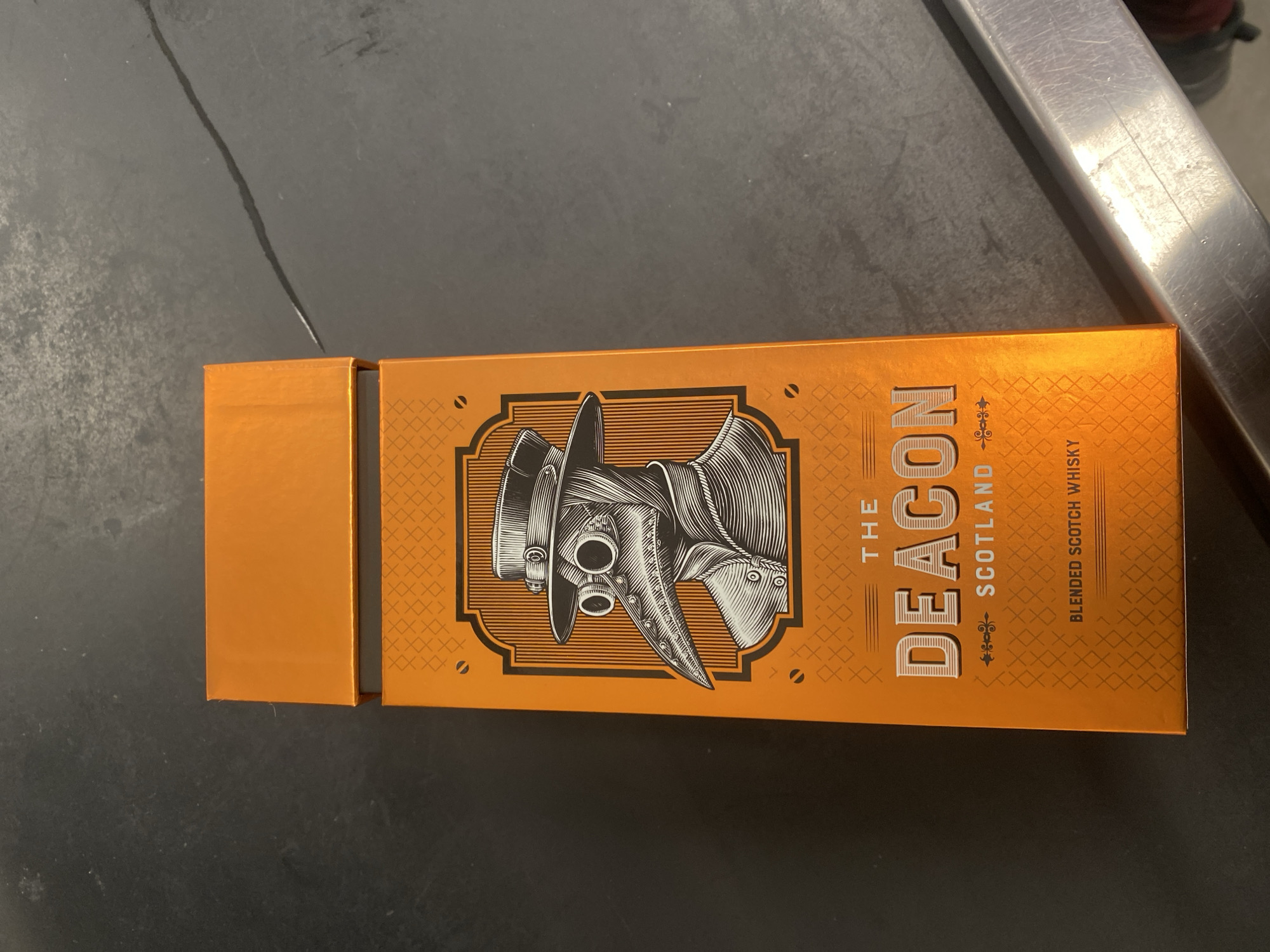 The image shows a box of "The Deacon" blended scotch whisky. The box is predominantly orange and features a black and white illustration of a plague doctor mask. "The Deacon" is printed vertically on the box. Below that is "Scotland" and "Blended Scotch Whisky." The box is on a dark surface, possibly a counter, next to a silver-colored object. The location of the image is unknown, and there's no information to suggest it is in Varna, Bulgaria. There are no people visible in the image.