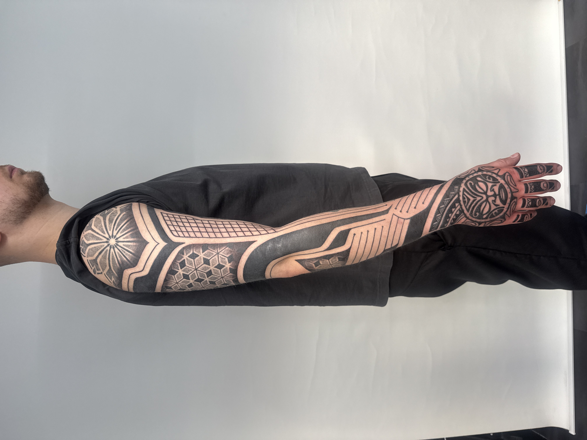 The image depicts a person lying down, presumably supine, with their right arm extended and prominently displaying a full sleeve tattoo. The tattoo features geometric patterns, including dot work, grid designs, and solid black areas. The tattoo extends onto the hand, covering the fingers with black ink and circular designs.
The person is wearing a dark t-shirt and dark pants. A portion of their face, including a beard, is visible. The background appears to be a plain white or light grey surface, likely a wall or a backdrop. The photograph has been rotated 90 degrees counter-clockwise.
There is no information in the image to indicate that it was taken in Sofia, Bulgaria.
