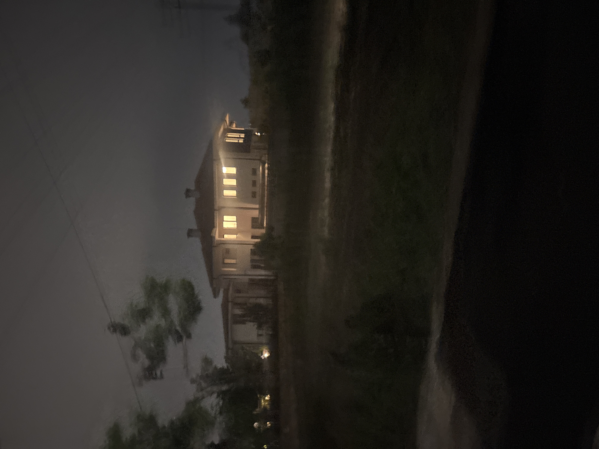 The image depicts a large building at night. The building appears to be multi-storied, with several illuminated windows visible. The architecture is characterized by a light-colored facade. To the right of the building is a dark area, potentially representing trees or other vegetation. The overall environment appears to be dim or dark, with a visible sky overhead. The presence of electrical wiring in the sky suggests a location within an urban or suburban area. The exact location in the city of Plovdiv, Bulgaria is indeterminable with the given information.