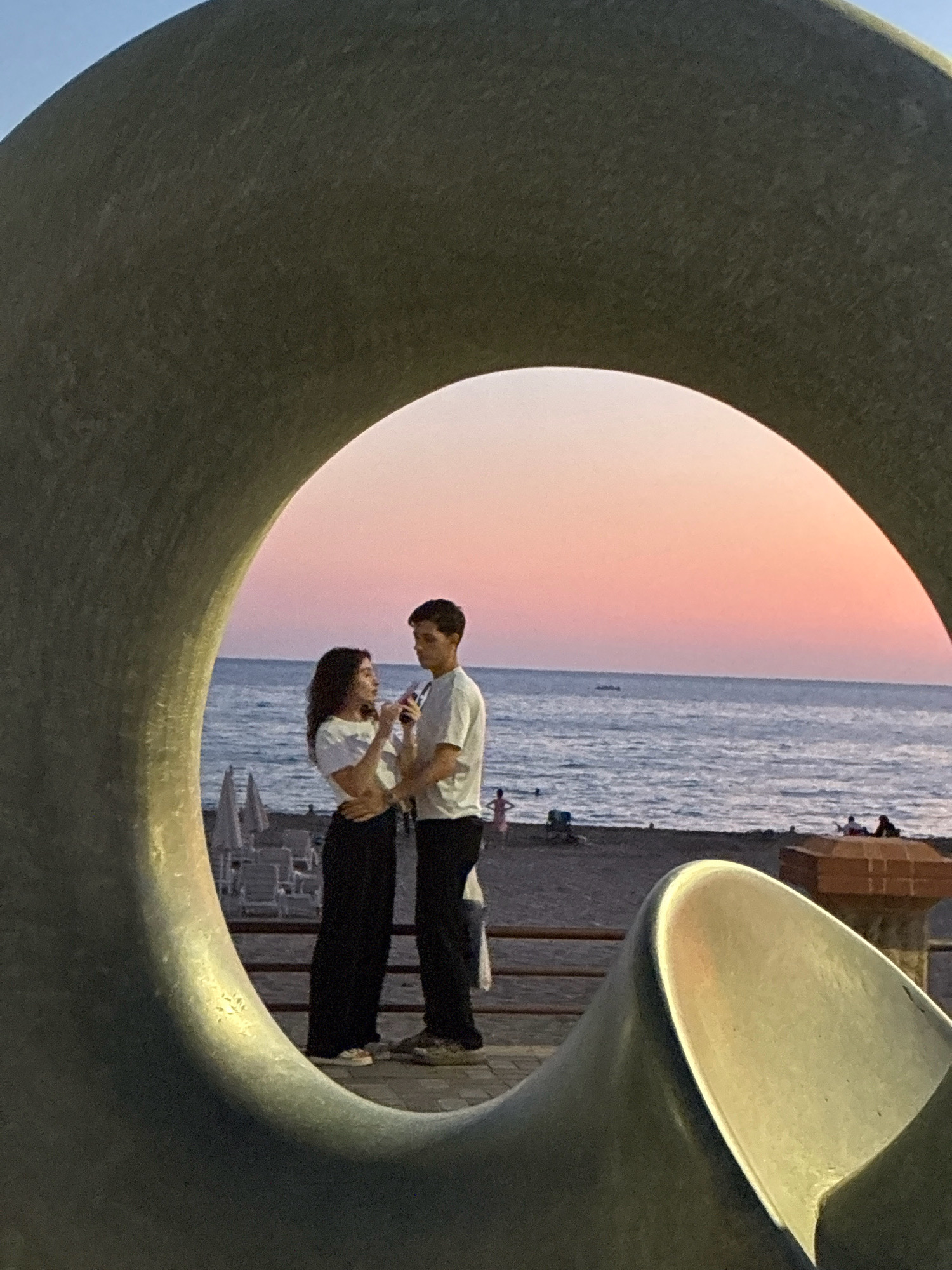 In Alanya, Turkey, a young couple stands close together, framed by a large, circular metallic sculpture. They are on a paved area with a railing overlooking a beach. The couple is facing each other, appearing to have a conversation or share a moment, with the woman touching the man's arm. Both are dressed casually in white tops and dark pants.
Behind them, the sea is visible, with gentle waves. The sky above the sea is painted with the soft hues of a sunset, transitioning from a light blue near the horizon to a pale pink. The beach is dotted with a few sunbathers and umbrellas, indicating it is a public space. The overall atmosphere is serene, capturing a moment of intimacy against the backdrop of a beautiful sunset.