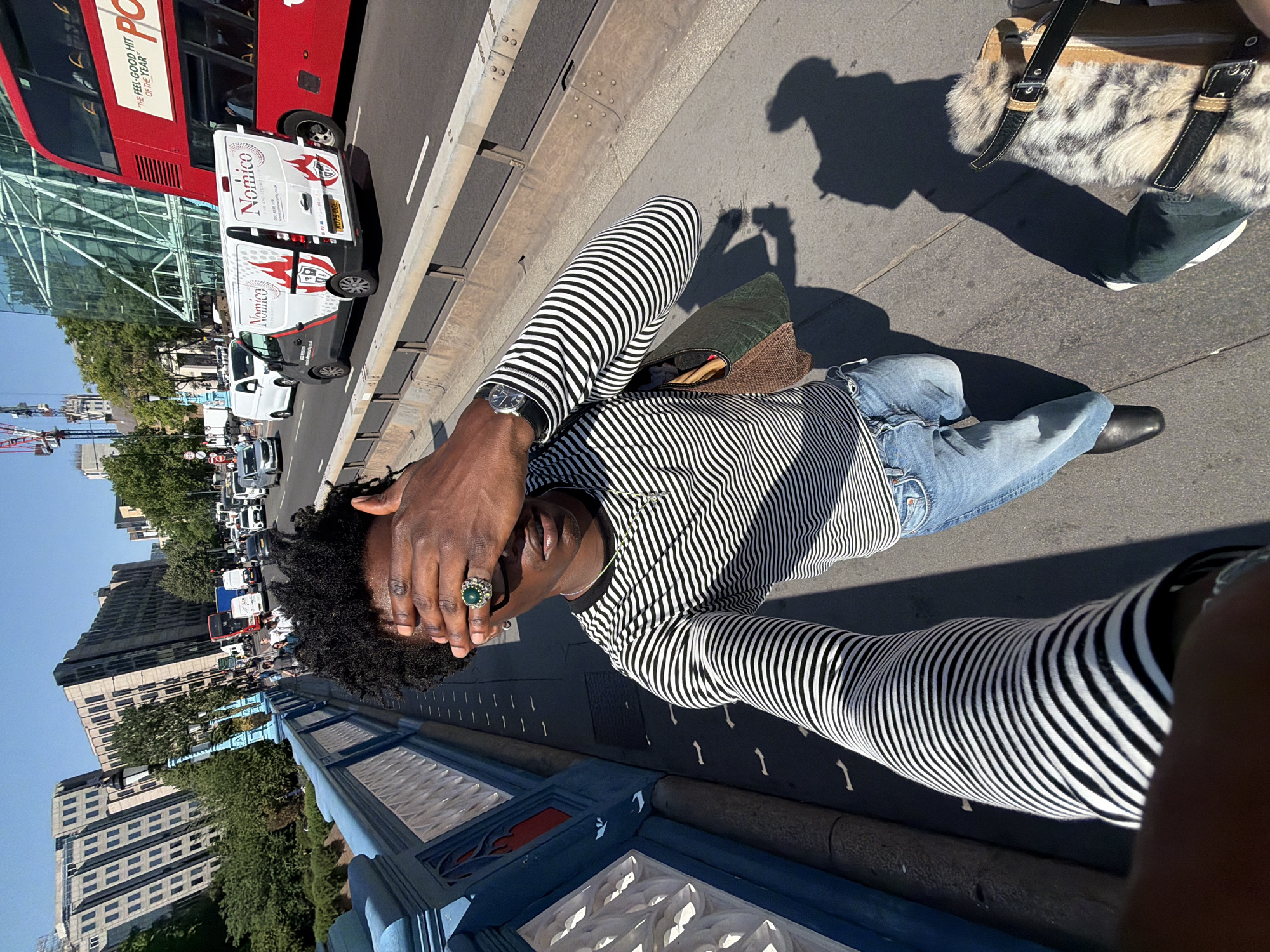 A person lies on a sidewalk next to a blue and red painted barrier. They are wearing a long-sleeved black and white striped shirt, blue jeans, and black shoes. The person has their hand covering their face and is wearing a large ring. Behind the person is a busy street with cars, vans with "Nomico" on the side, and a red double-decker bus. A building with scaffolding around it is also visible. In the foreground to the right is part of a beige and black patterned bag. The image appears to have been taken on the Tower Bridge in London, United Kingdom.
