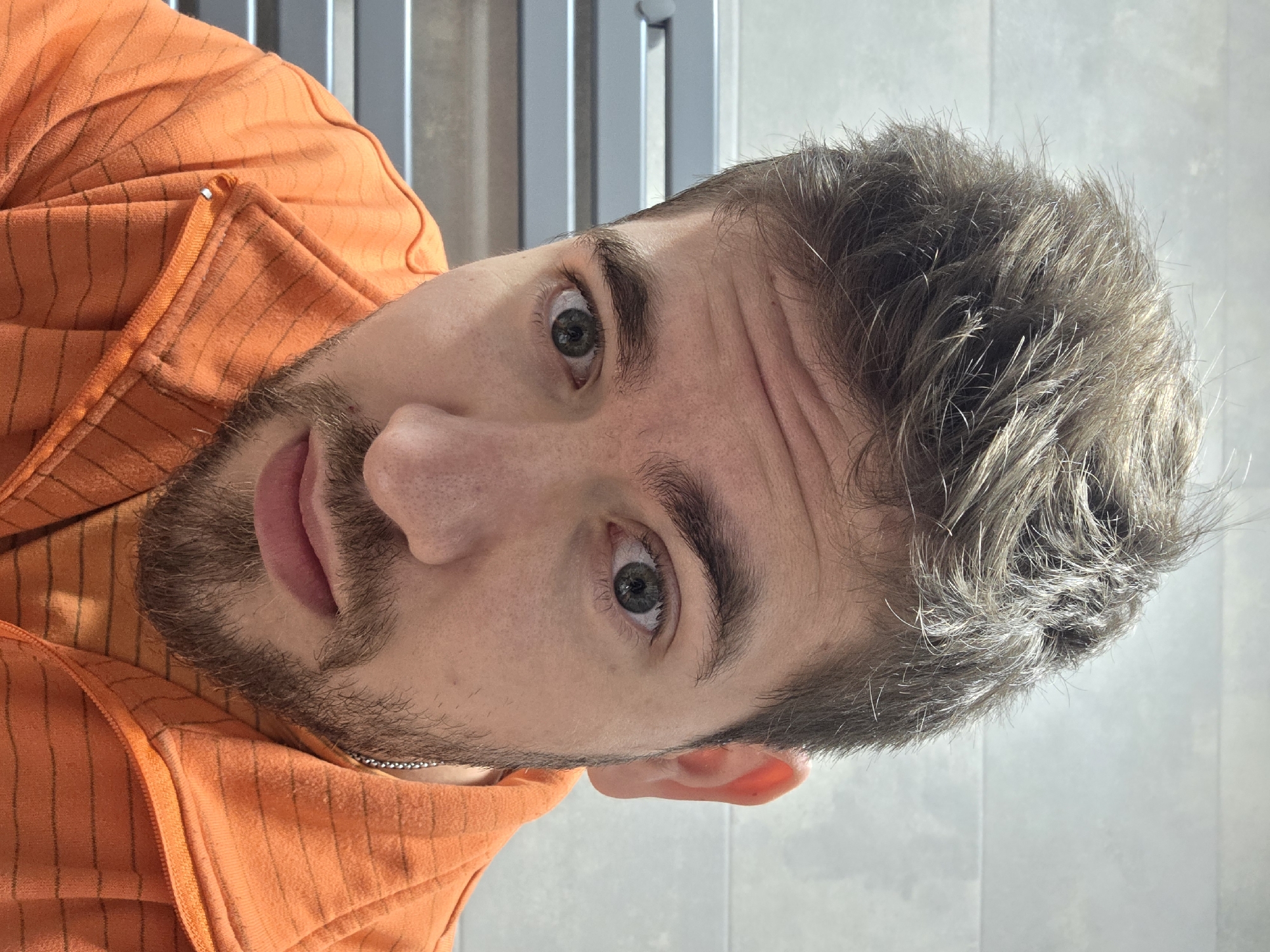 A man is seen from a close-up, high-angle perspective. He is wearing an orange hooded sweatshirt. The man has short, grey-brown hair and a beard. His eyes are green. A grey, metal radiator and a light grey tiled wall are visible in the background. The location is unknown, so no reference to Yagodovo, Bulgaria can be made.