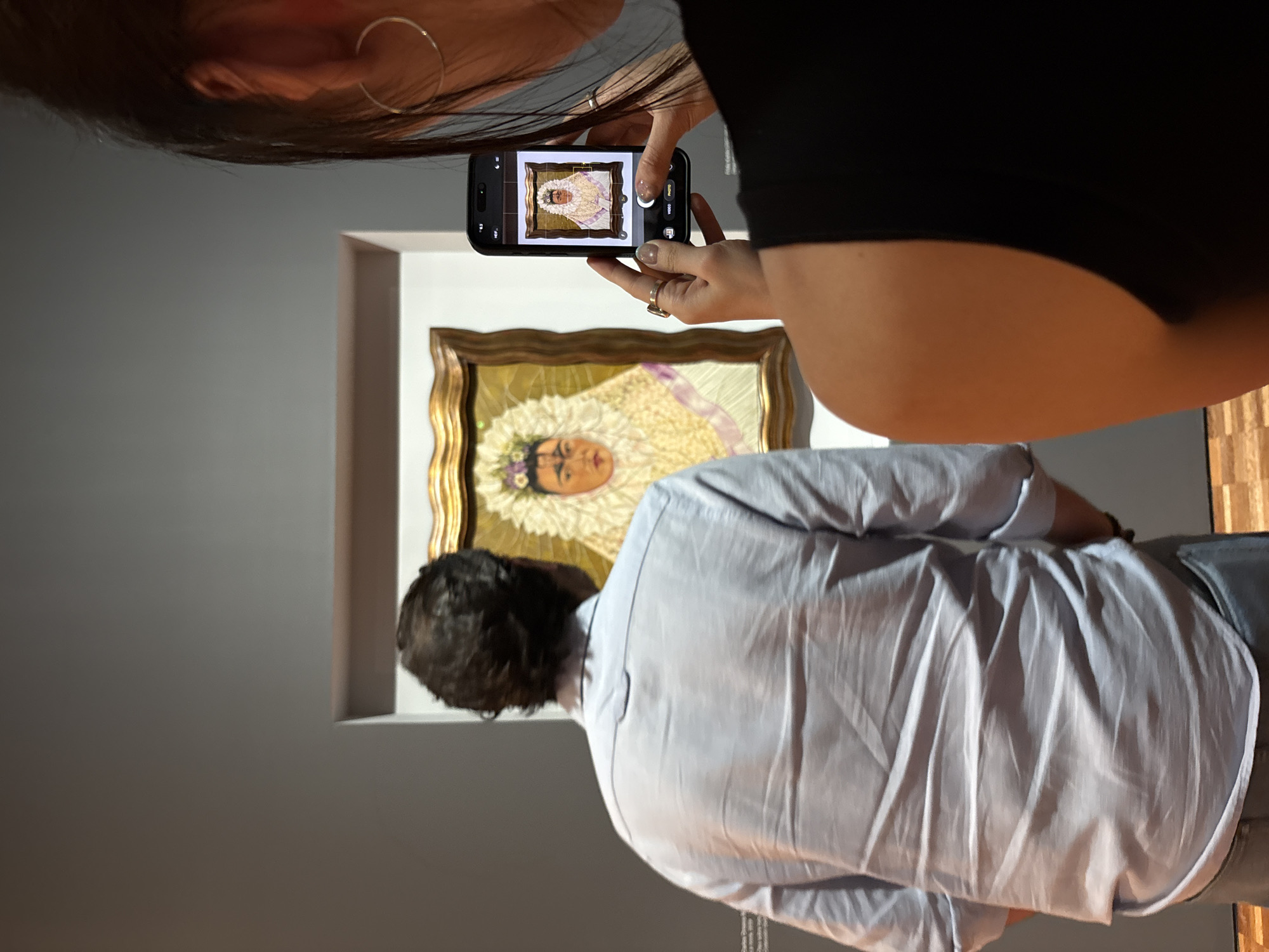 A woman is taking a picture of a portrait with her phone. The portrait, featuring a woman with flowers in her hair, is framed in a decorative gold frame and hangs on a white wall. A man is standing in front of the portrait, viewing the painting. The setting appears to be inside a museum or art gallery. The painting is a portrait of Frida Kahlo, indicating that the location is most likely in Mexico City, Mexico, where Frida Kahlo spent much of her life and where her work is prominently displayed.