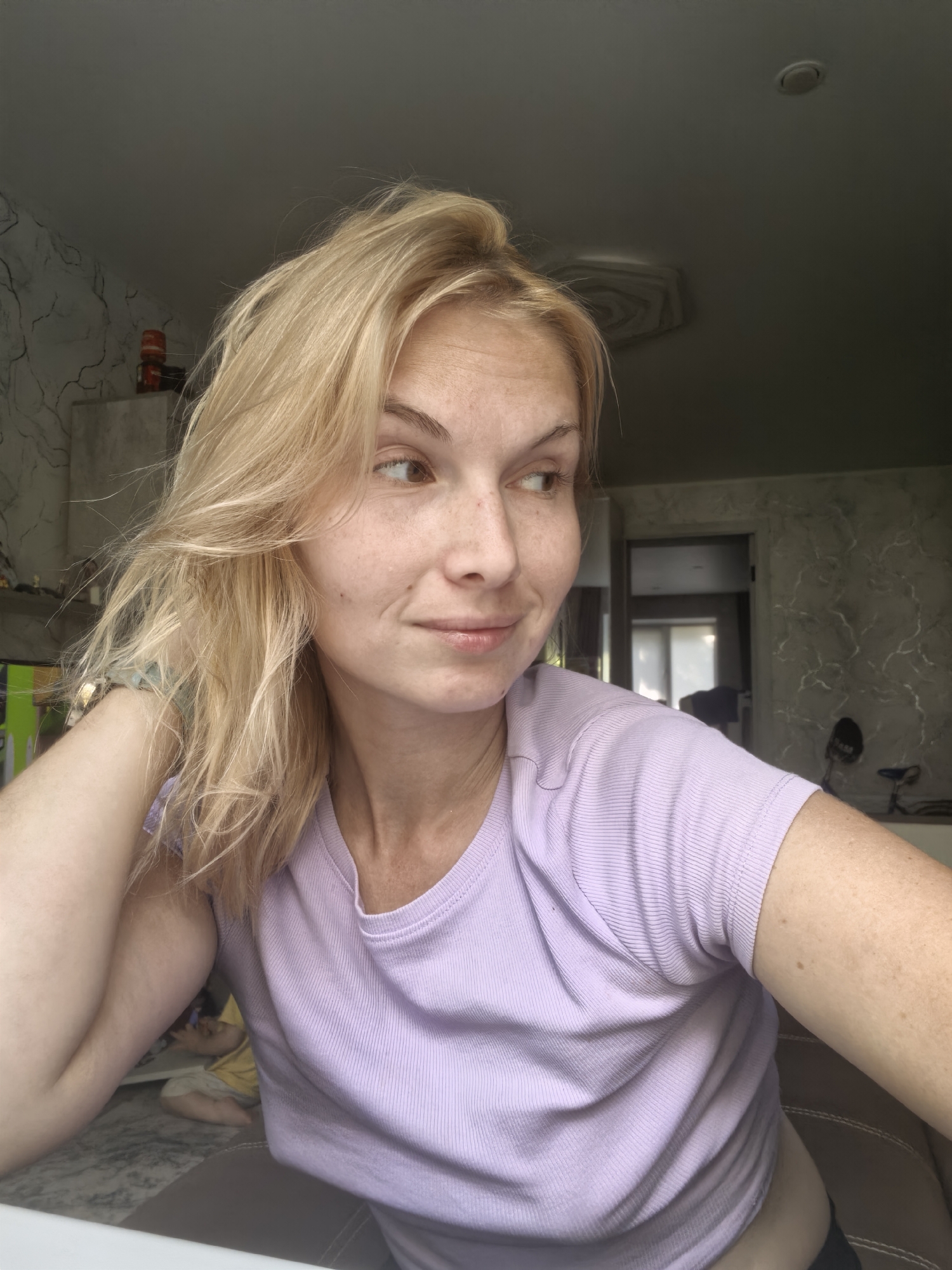 A woman with blonde hair and freckles is captured in a medium shot, looking to her right. She is wearing a light purple, ribbed t-shirt. Her left arm is bent, with her hand near her head, and her right arm is resting on her side. The background shows an indoor setting, possibly a room in an apartment in Sterlitamak, Russia. A doorway and part of a wall with a patterned texture are visible behind her. In the blurred background to the left, a person appears to be lying down on a couch or bed.