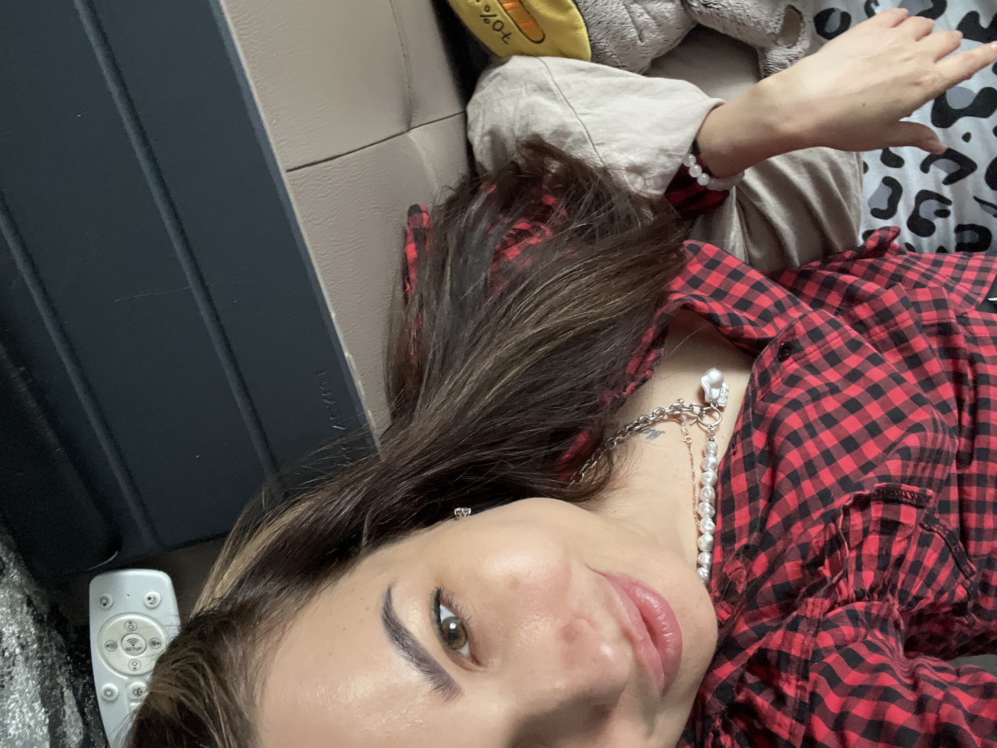A close-up, low-angle shot shows a woman lying on a surface, possibly a couch or bed, in an indoor setting. The location appears to be in an unidentified city, province, or state.
The woman has dark hair with highlights, visible skin, and makeup on her face. She's wearing a red and black plaid shirt and a pearl necklace. A white remote control is visible near her head.
In the background, there are items such as a tan-colored blanket or pillow and a black and white patterned sheet or blanket. A dark gray or black object, possibly a laptop case or a piece of furniture, is positioned behind her head. The lighting is soft, suggesting natural or diffused light.