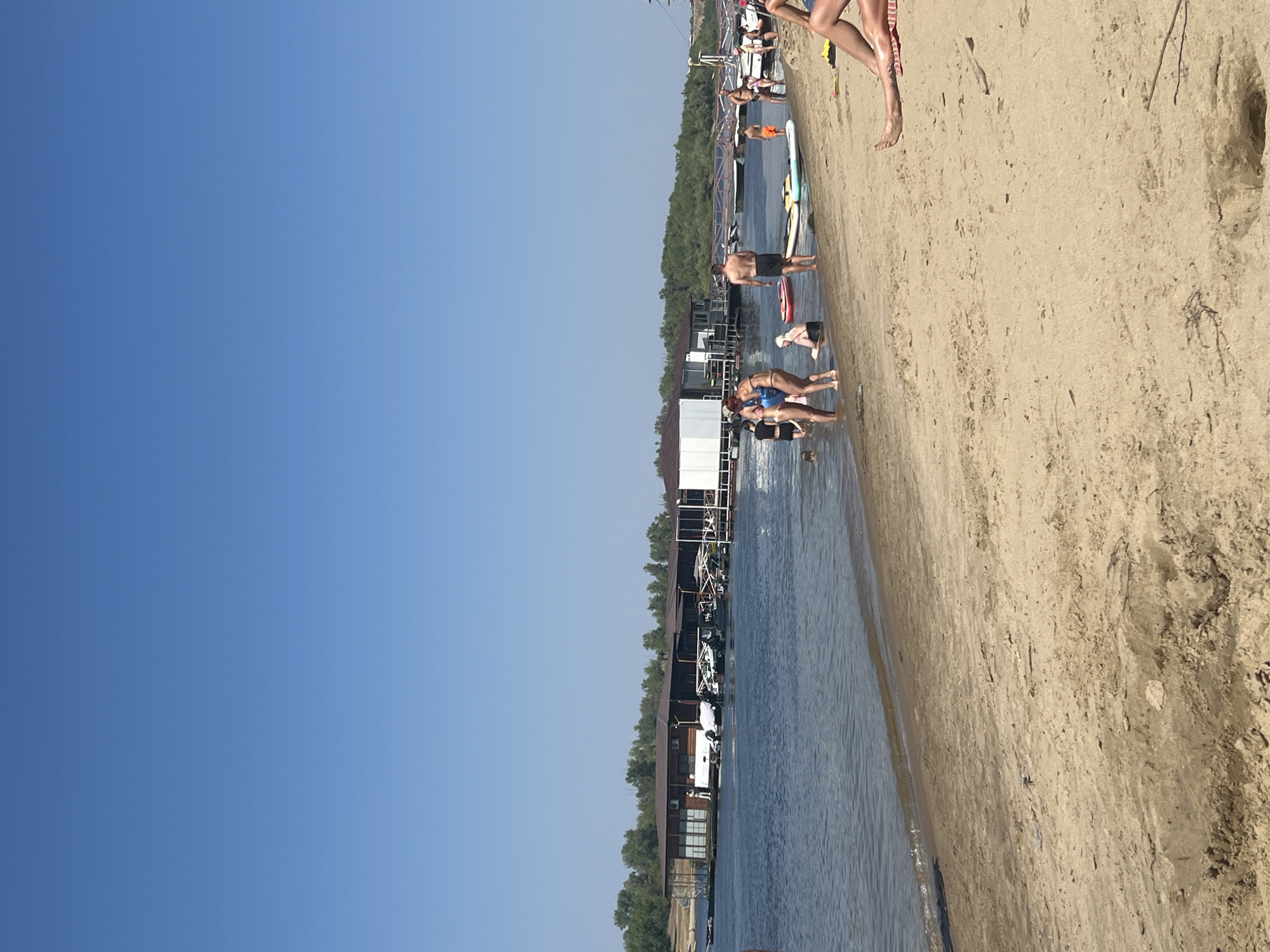 A wide shot captures a sunny day on the sandy beach of Kilyakovka, Russia. The scene is bustling with activity as people enjoy the water and the shore. In the distance, a series of large, wooden, dock-like structures with white roofs and wooden supports extend over the water. Several boats are moored to these structures, and a few people can be seen on them. The water is calm and reflects the clear blue sky. Closer to the shore, several individuals are wading in the shallow water, with some appearing to be children. On the sand, a few people are standing or sitting, while others are lounging on towels. In the foreground, the sandy beach stretches out, with scattered debris such as small branches and seaweed. The overall impression is a lively and enjoyable summer day by the water.