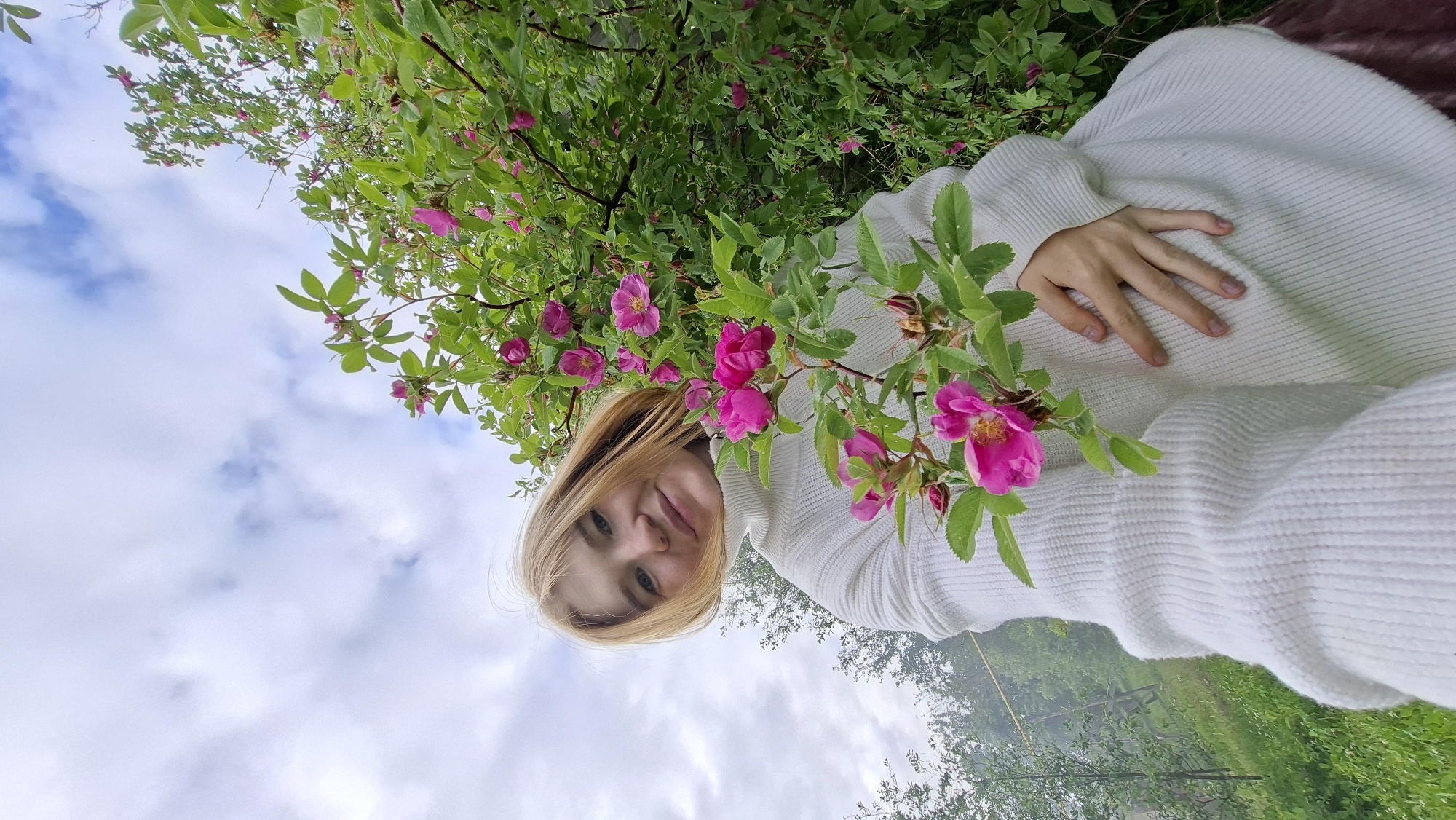 A young Caucasian woman with blonde hair and fair skin is pictured from a low-angle perspective, looking towards the camera. She is wearing a white, high-necked, chunky knit sweater. Her right hand is placed on her chest, partially obscured by a branch of pink flowers. The flowers are in bloom on a lush green bush behind and to the right of her. Her expression is a slight smile. The background consists of a slightly misty, green landscape with trees and vegetation, and a cloudy, overcast sky. The location is identified as Большой Букор, Russia.
