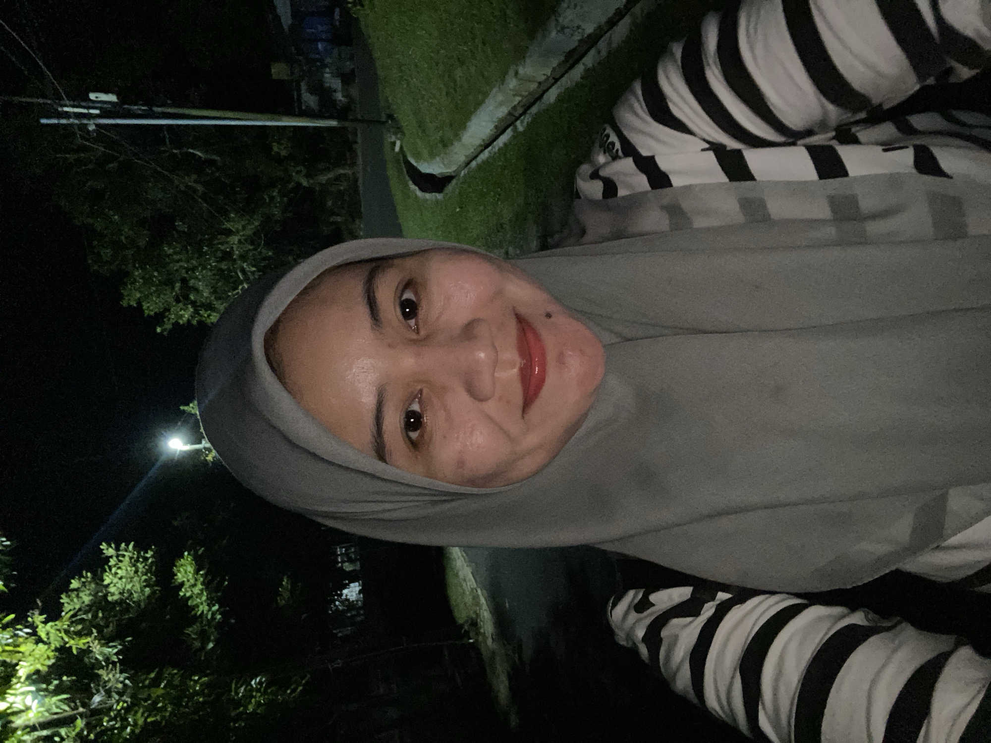 A medium shot shows a woman wearing a gray hijab and a black and white striped shirt. She is standing outdoors at night. The background includes trees, a street, a grassy area with a drainage ditch, and a utility pole. The scene appears to be in a residential or park area within the city of Banjarbaru, Indonesia, given the tropical vegetation. The lighting is dim, with a streetlamp illuminating part of the area.