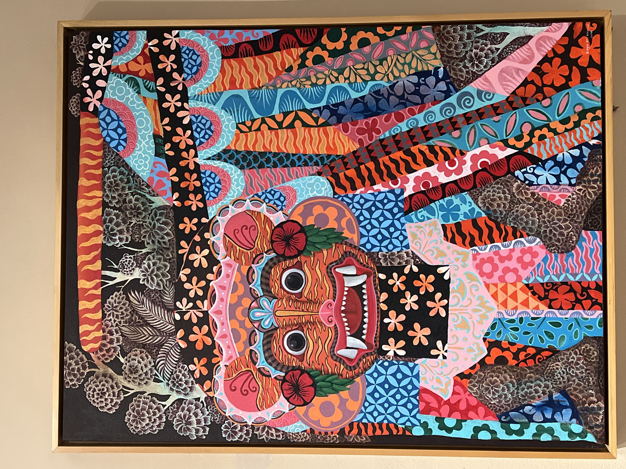 This is a detailed, vibrant painting, presented within a light wood frame, depicting a stylized mask-like face and intricate patterns, located in Ubud District, Indonesia. The central figure is a large, orange and black striped face, resembling a tiger or mythical creature, featuring prominent white fangs, a red mouth, a small pink nose, and large, round black eyes with white outlines. Red floral elements with green leaves adorn the sides of the face, which is topped with a pink and light blue crown-like headpiece.
The background and body of the figure are composed of numerous distinct, colorful patterns, creating a patchwork or mosaic effect. These patterns include a variety of geometric shapes, floral motifs (such as orange flowers on a black background, pink flowers, and stylized white flowers), wavy lines, scales, and abstract designs. Dominant colors in these patterns include various shades of blue, pink, red, orange, green, black, and yellow.
In the upper section, against a black background, are stylized trees and foliage rendered in muted grays, whites, and subtle greens. A horizontal orange and yellow striped band is visible at the very top. The right side of the painting features a dynamic cascade of vertically oriented, fan-shaped and wavy strips filled with diverse patterns. The lower sections of the painting also continue this intricate patchwork of colorful designs, interspersed with darker, textured forms that resemble stylized foliage or ground elements. The painting is framed with a simple wooden border and hangs on a plain, light-colored wall.