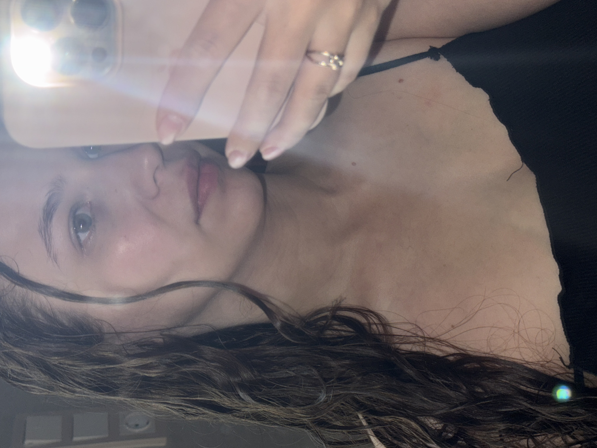 The image is a close-up, selfie-style photograph of a young woman's face and upper body. She has dark, wet, curly hair that falls over her left shoulder. Her face is turned towards the camera, and she is holding a light pink smartphone in her right hand, which is partially obscuring her face. The phone's flash is activated, creating a bright glare in the image. She is wearing a black spaghetti-strap top. A ring with multiple small stones is visible on her right ring finger. The background is blurred and appears to be indoors, possibly a bathroom or bedroom. There are some indistinct objects on the wall to the left, one of which resembles a circular clock. The lighting is harsh due to the phone's flash. There is no information in the image to suggest it is located in Chaikovsky, Russia.
