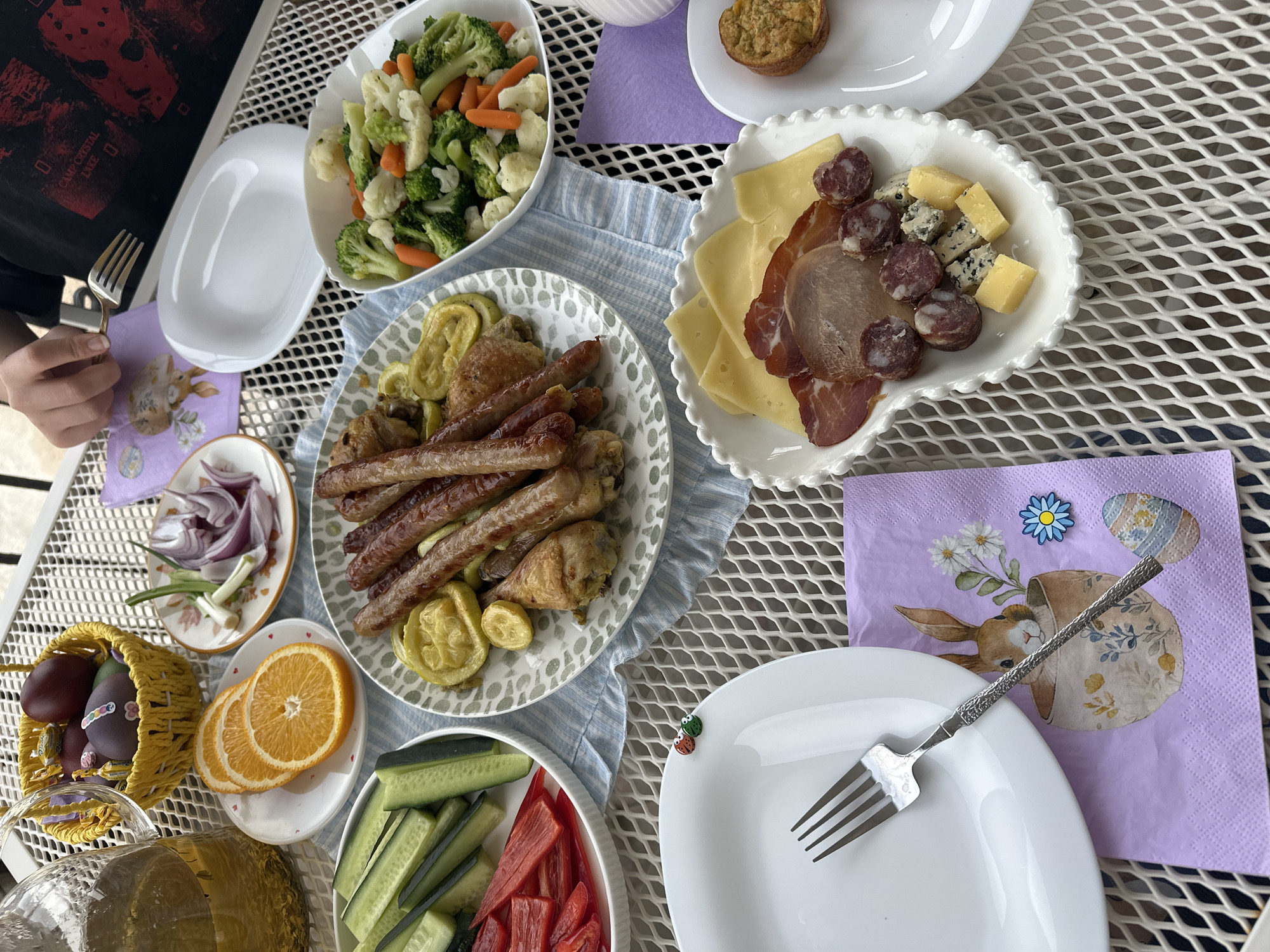The image depicts an outdoor table setting, laden with food, likely for a meal or gathering.
**Subjects:** The primary subject is the food and the table arrangement. A person's hand holding a fork is visible in the top left corner, suggesting an intention to eat.
**Objects:** The objects present are varied and numerous, including:
* Plates of different sizes and shapes, some with decorative edges.
* Food items: assorted cooked sausages, chicken pieces, and zucchini on one plate; a bowl of mixed steamed vegetables (broccoli, cauliflower, carrots); a charcuterie board with cheese, cured meats, and sausage; slices of orange and cucumber; sliced red onion.
* Tablecloths/napkins, one light blue striped and others with Easter-themed patterns.
* Forks with distinctive handles.
* Easter eggs in a basket.
* Metal table surface.
**Environment:** The setting is outdoors, suggested by the metal surface with a grid pattern, resembling patio furniture.
**Actions:** The action in progress is the preparation for or enjoyment of a meal.
The image does not show location information related to Bar, Montenegro.