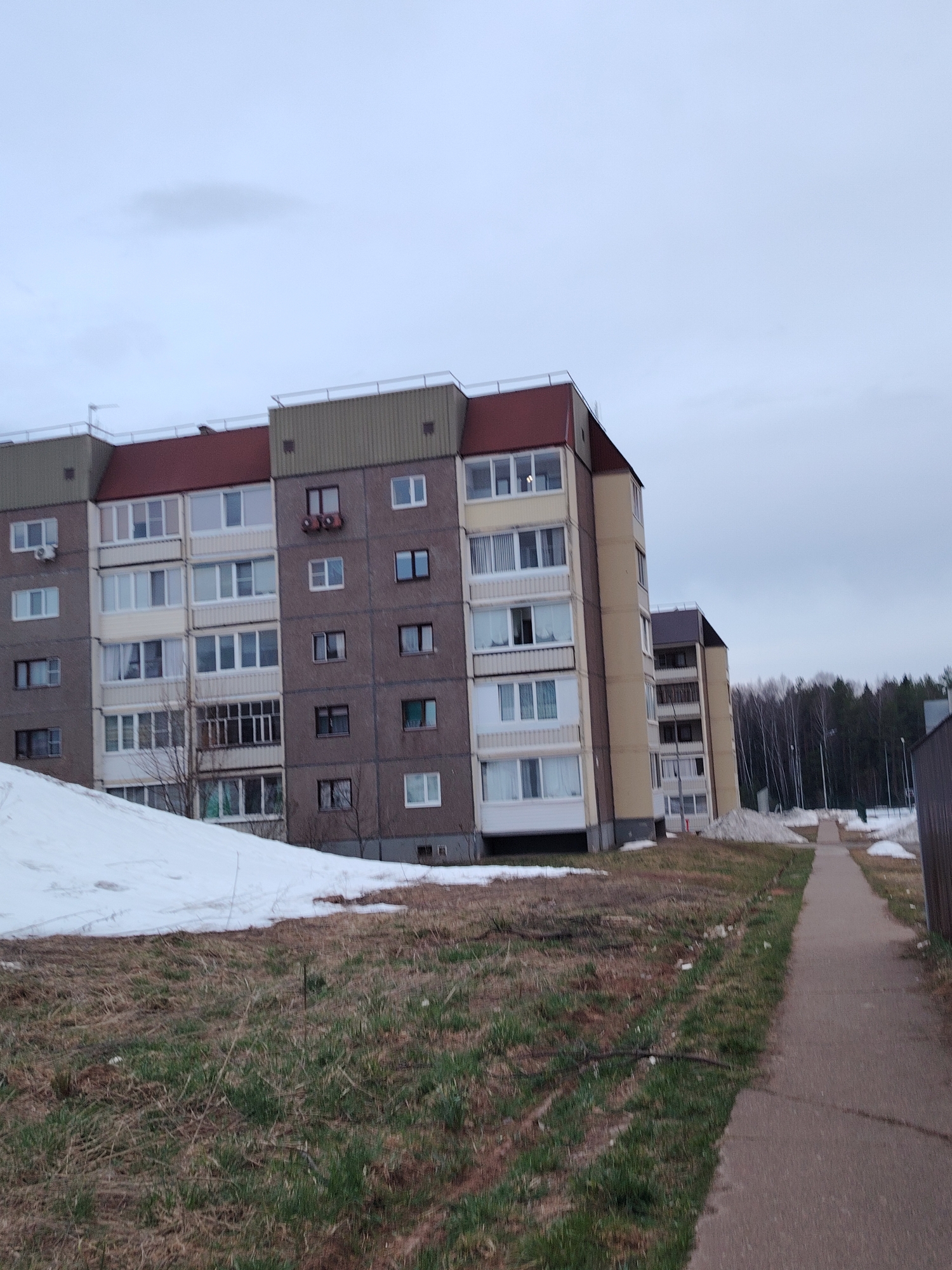 The image shows an apartment building in Марковский, Russia. The building is a multi-story structure with a mix of concrete and painted facades. There are multiple windows on each level. Some of the windows have balconies. The ground is covered with a mix of grass, dirt, and patches of snow. A sidewalk runs along the side of the building. In the background, there is a forest and another apartment building. The sky is overcast.