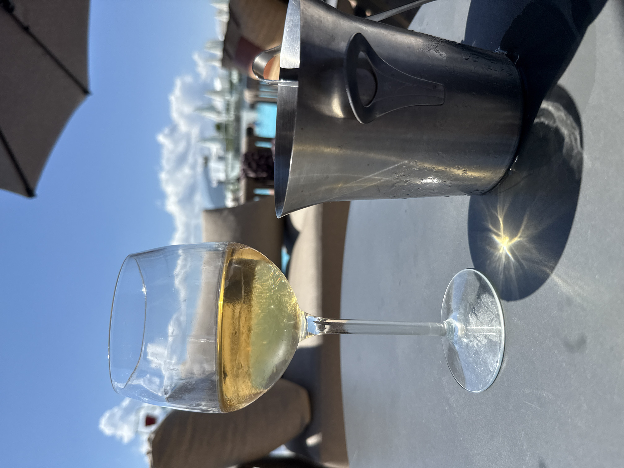 The image depicts an outdoor leisure setting under a clear blue sky with scattered white clouds, located in the city of Unknown, Unknown. In the foreground, a stemmed wine glass, approximately half-full with a light yellow liquid (white wine), stands on a dark grey, textured table surface. To the right of the wine glass, a cylindrical, stainless steel ice bucket with ice tongs inserted is also present on the table. The ice bucket shows condensation and water droplets on its exterior, indicating it contains ice. Strong direct sunlight from the upper left casts distinct shadows of both the wine glass and the ice bucket onto the table. A bright, starburst-shaped sun glare is visible on the table surface to the right of the wine glass's base and extending into the ice bucket's shadow.
In the mid-ground, out of focus, are several upholstered lounge chairs or daybeds with beige cushions. Behind these chairs, a body of water, likely a swimming pool, is visible, along with multiple white, architectural or decorative structures that appear somewhat uniform in design. A dark-colored patio umbrella is partially visible in the upper right corner of the frame. To the far left in the background, partially obscured by a lounge chair, a faint object resembling a glass with a red liquid can be discerned. No people are visible in the scene. The overall lighting suggests a sunny daytime environment.