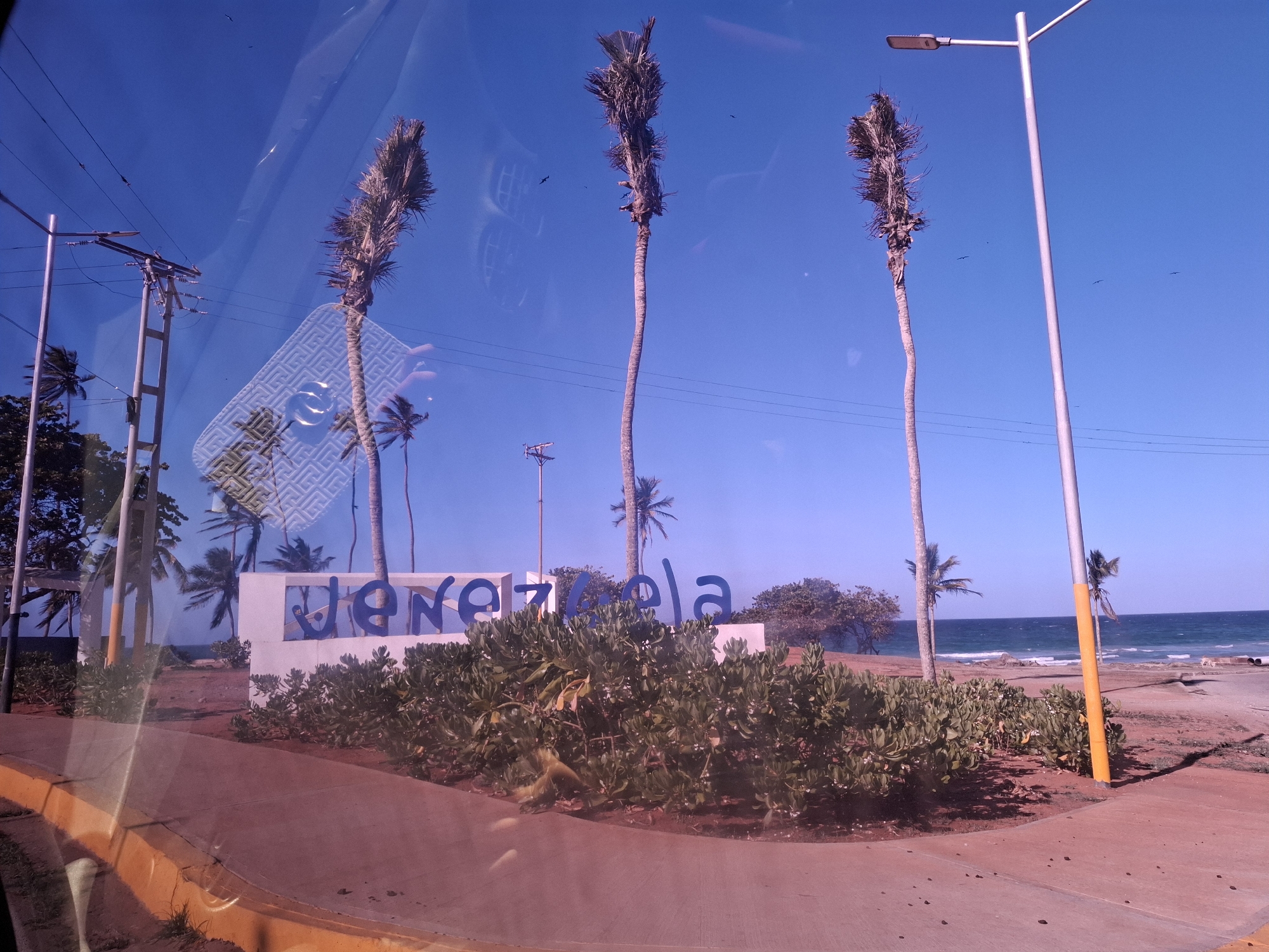 This media file captures a bright, sunny coastal landscape, likely taken during the daytime from inside a vehicle due to reflections visible on what appears to be a window. The scene is dominated by a clear, vibrant blue sky.
In the foreground, a paved path or road with a distinct yellow curb is visible. A bed of lush green bushes separates the path from a prominent white sign featuring large, blue, three-dimensional letters that spell out "Venezuela."
Behind the sign and foliage, three tall, slender palm trees with somewhat sparse fronds stand against the sky. To the left, utility poles with wires are visible, while a modern streetlight post extends upwards on the right. More distant, smaller palm trees are scattered throughout the background.
Beyond the land features, the deep blue waters of the ocean stretch out, with gentle waves breaking on the shore, suggesting a beach or rocky coastline. Several small, dark bird silhouettes are visible against the expansive blue sky. The overall scene evokes a calm, tropical coastal atmosphere. No people or specific activities are depicted. The visible text clearly identifies the location as "Venezuela."