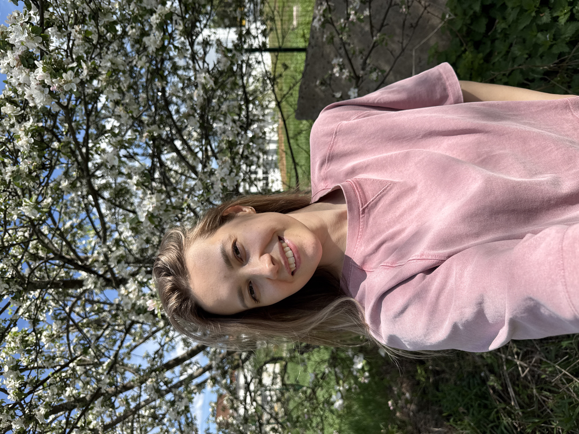 A woman wearing a pink sweatshirt is smiling and looking towards the camera. She stands outside near a flowering tree. A fence and grassy area are visible in the background. The location is Noviy, Russia.