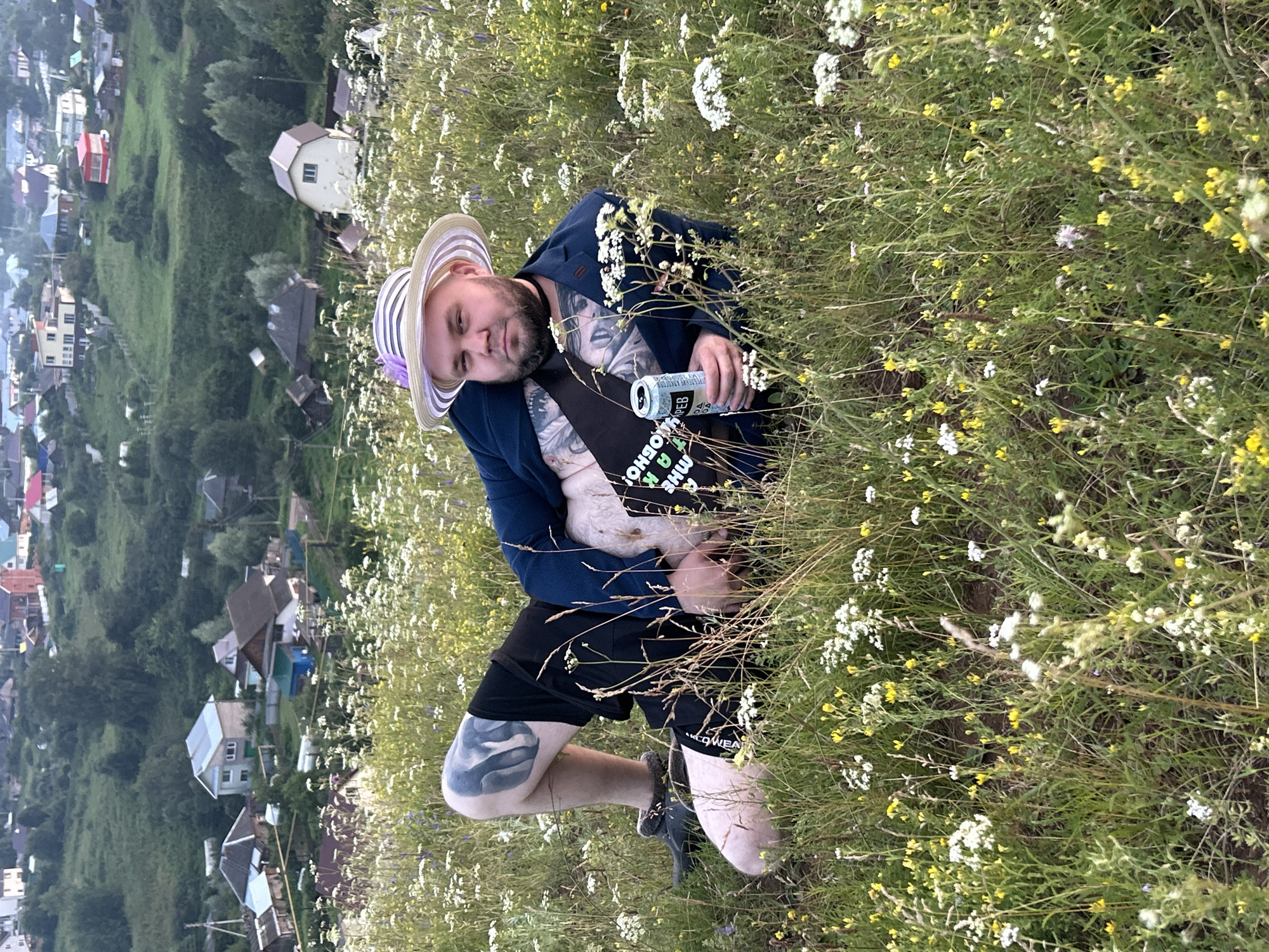 In the city of Olkhovka, Russia, a man with a beard and tattoos is lying in a field of wildflowers. He is wearing a dark blue blazer, black shorts, and a striped hat. A can of beer is in his hand, and a sash with Cyrillic text is draped across his chest. The background shows a hilly landscape with houses and trees. The lighting suggests it is late afternoon or early evening.